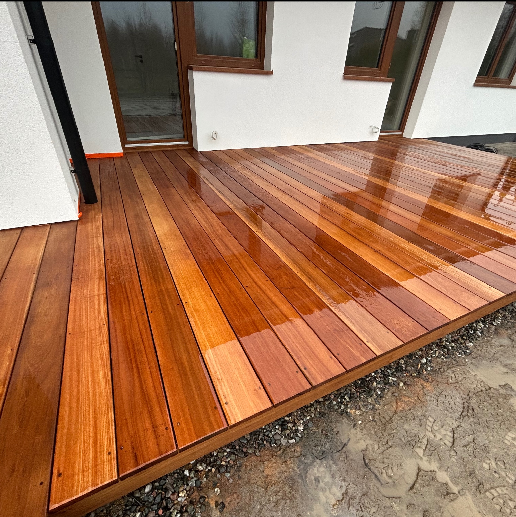 Świeżo zaimpregnowany taras z drewna egzotycznego, widoczne refleksy światła na mokrej powierzchni, obrzeże z drobnego kamienia, tło stanowią ściany budynku z oknami.