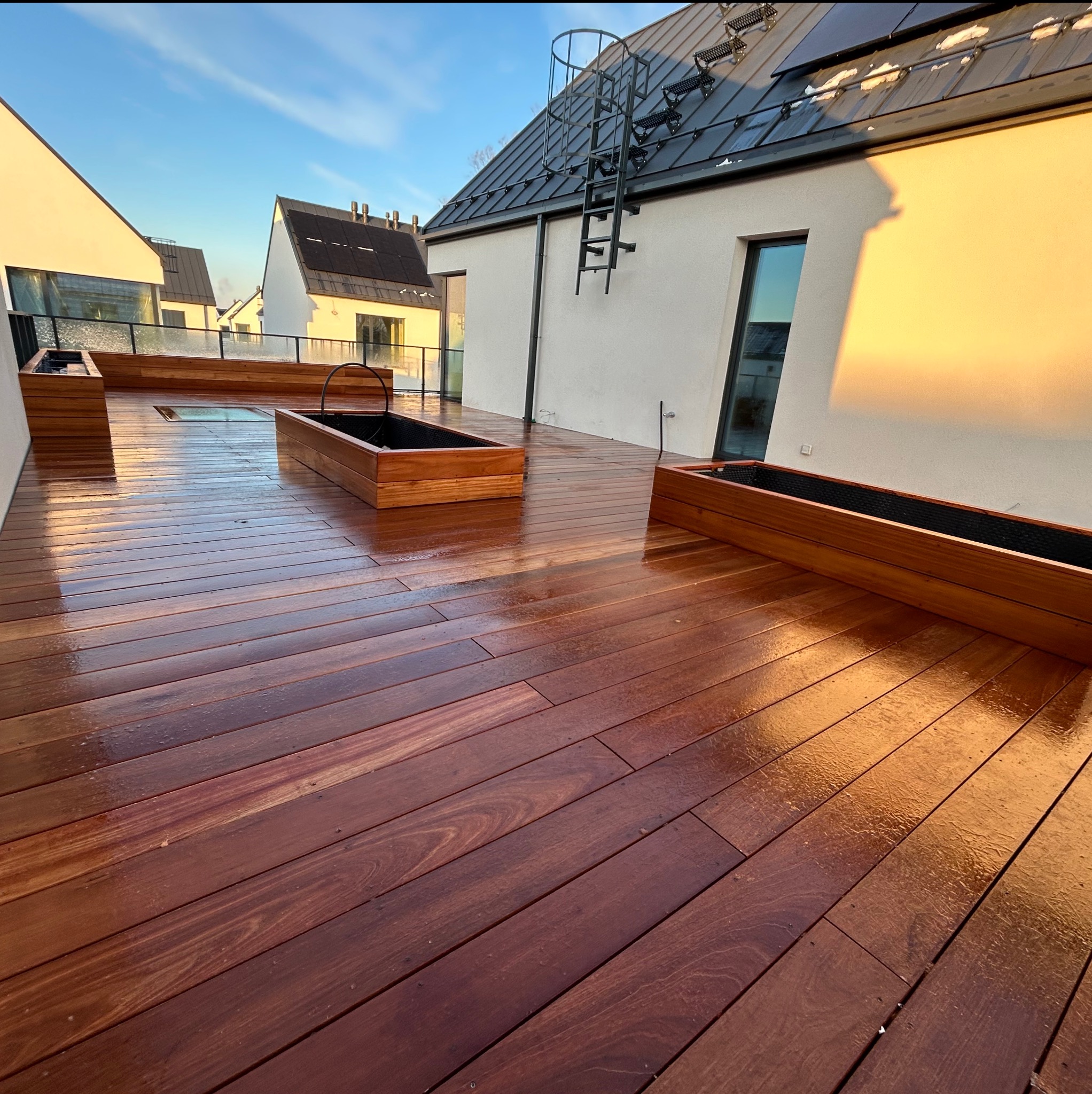 Błyszczący, świeżo wyczyszczony taras z ciemnego drewna z drewnianymi donicami, widok na nowoczesne domy w tle, wczesne popołudnie.