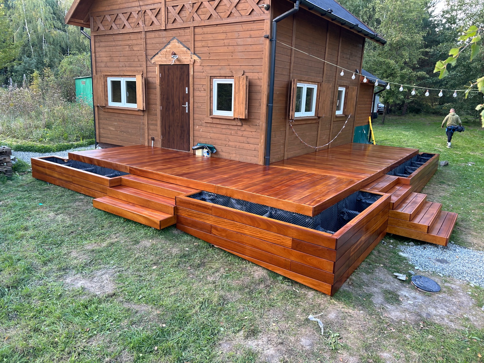 Nowoczesny taras z drewna egzotycznego, z wbudowanymi donicami, prowadzący do drewnianego domku, oświetlony girlandą żarówek, na tle zieleni drzew.