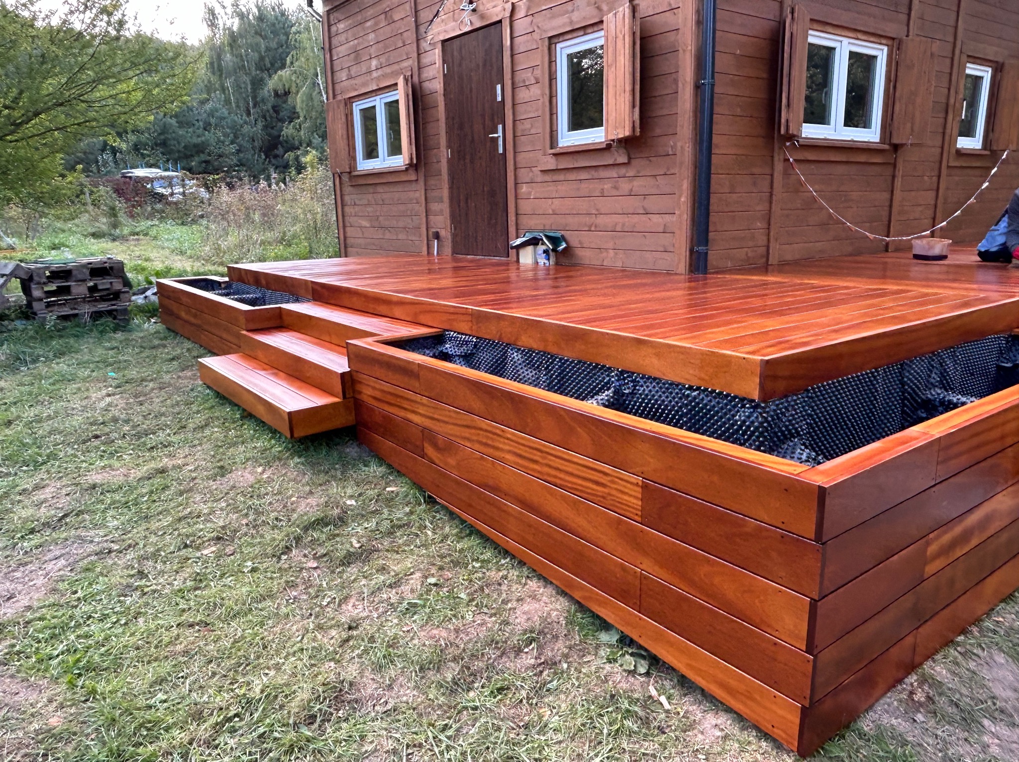 Błyszczący, drewniany taras z trzema stopniami prowadzącymi do domu z drewnianą elewacją i jasnymi ramami okiennymi. Taras posiada wbudowane skrzynie na rośliny z czarną wyściółką.