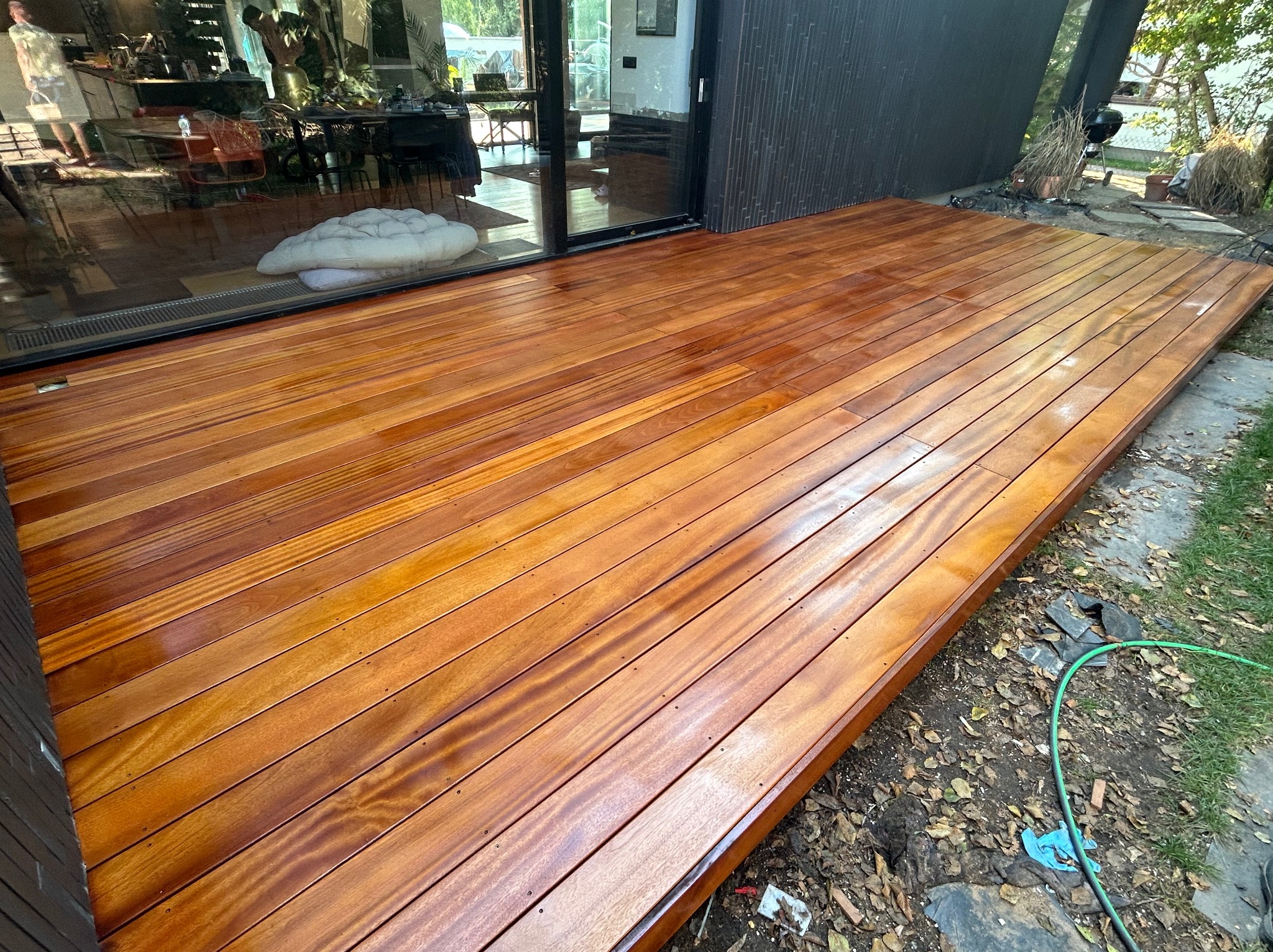 Świeżo polakierowany, brązowy taras z desek z widocznym odbiciem wnętrza domu w szklanych drzwiach tarasowych. Na zewnątrz widoczny fragment trawnika i wąż ogrodowy.