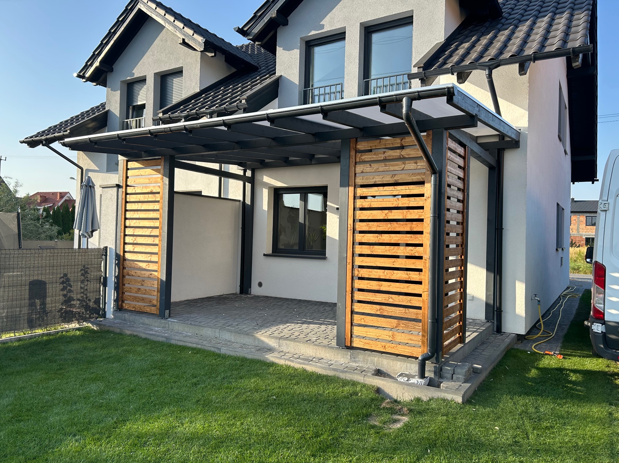 Nowoczesna pergola z drewnianymi lamelami na betonowym tarasie przy białym domu z ciemną dachówką, widok z poziomu trawnika.