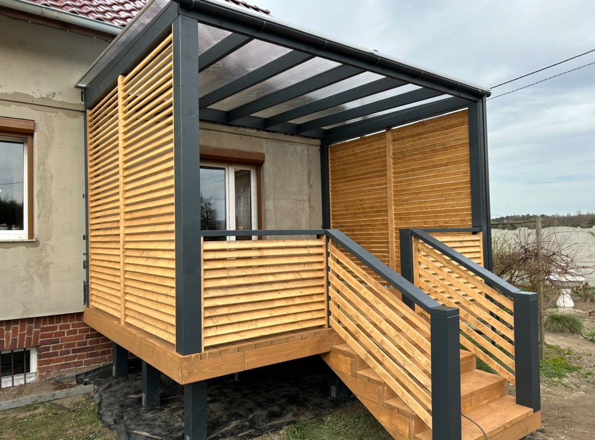 Zadaszona drewniana konstrukcja z żaluzjowymi ścianami, schodami i szarą ramą, przylegająca do budynku z oknami.