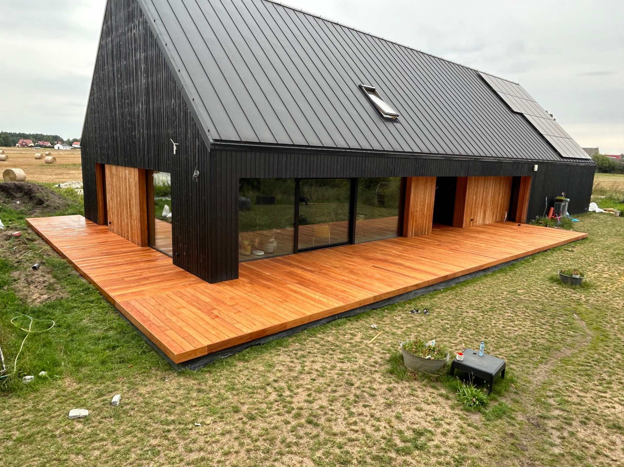 Nowoczesny dom z czarną elewacją i rozległym tarasem z drewna egzotycznego, widok z góry na trawnik z dekoracjami ogrodowymi i bale siana w tle.