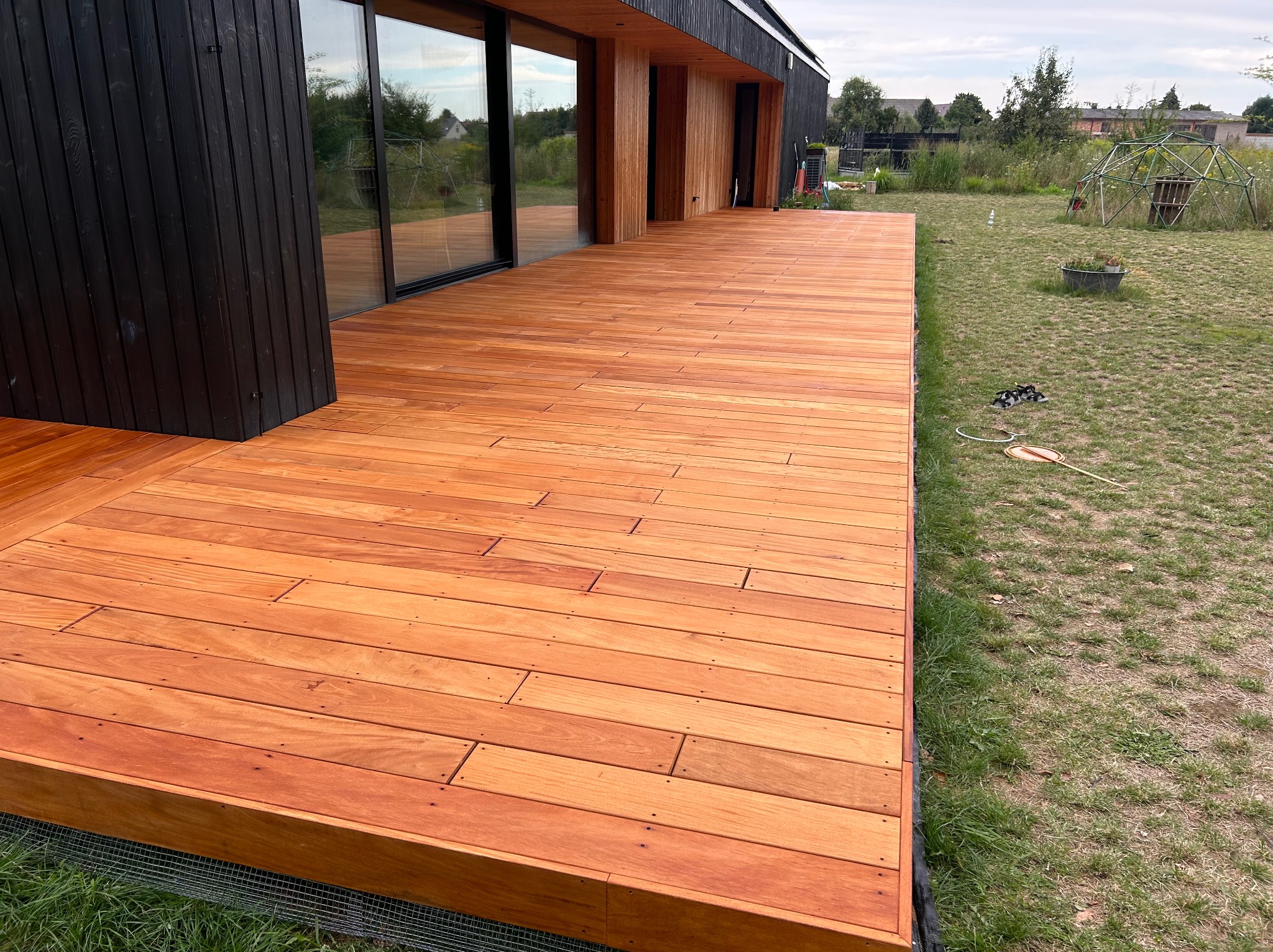 Nowoczesny dom z dużym tarasem z desek o ciepłym, pomarańczowo-brązowym odcieniu, przylegającym do czarnej elewacji. W tle widoczny trawnik z konstrukcją geodezyjną.