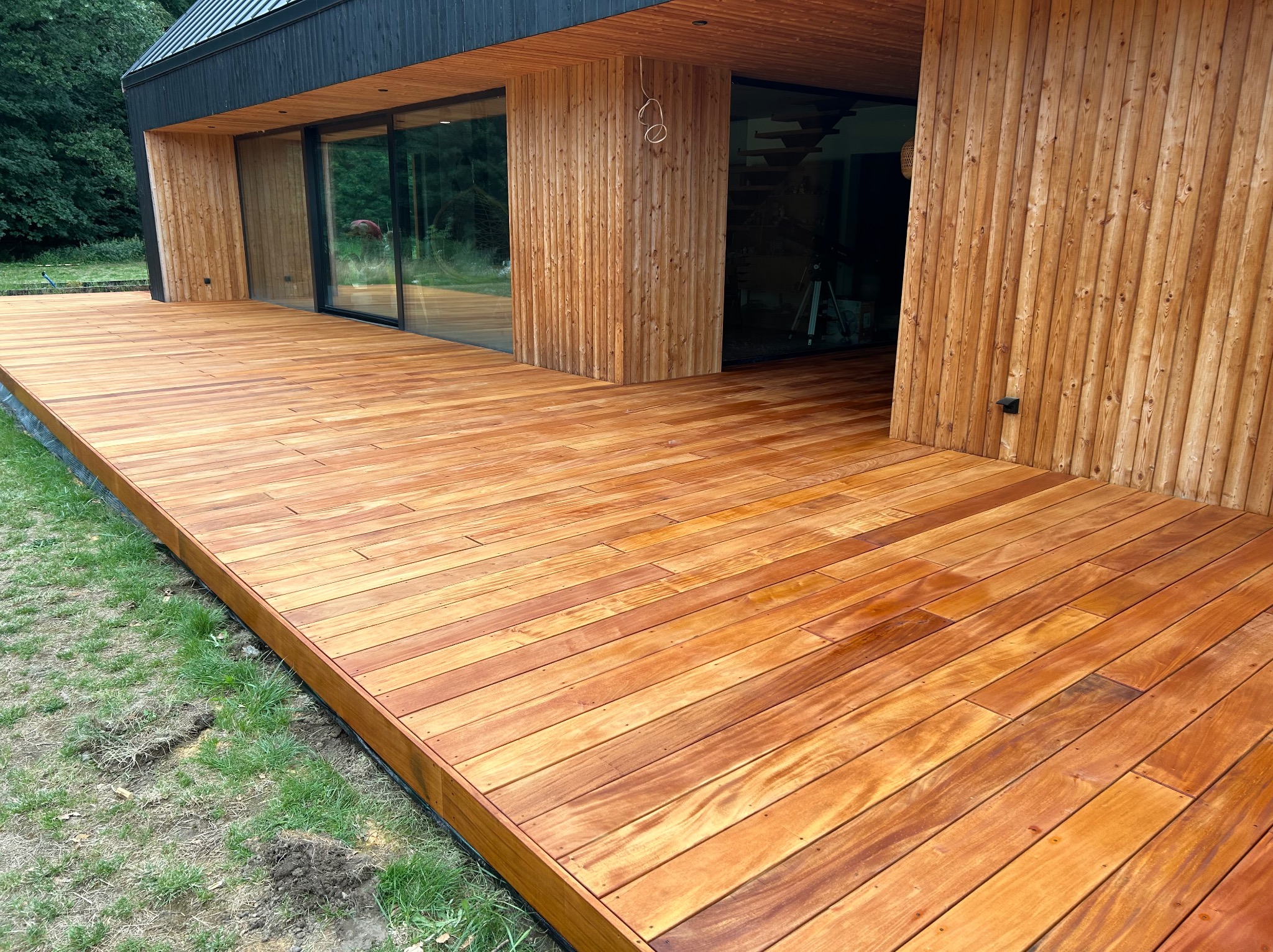 Nowoczesny dom z elewacją z jasnego drewna i dużym, drewnianym tarasem, widok z boku na świeżo położoną deskę tarasową, z widocznym trawnikiem na skraju.