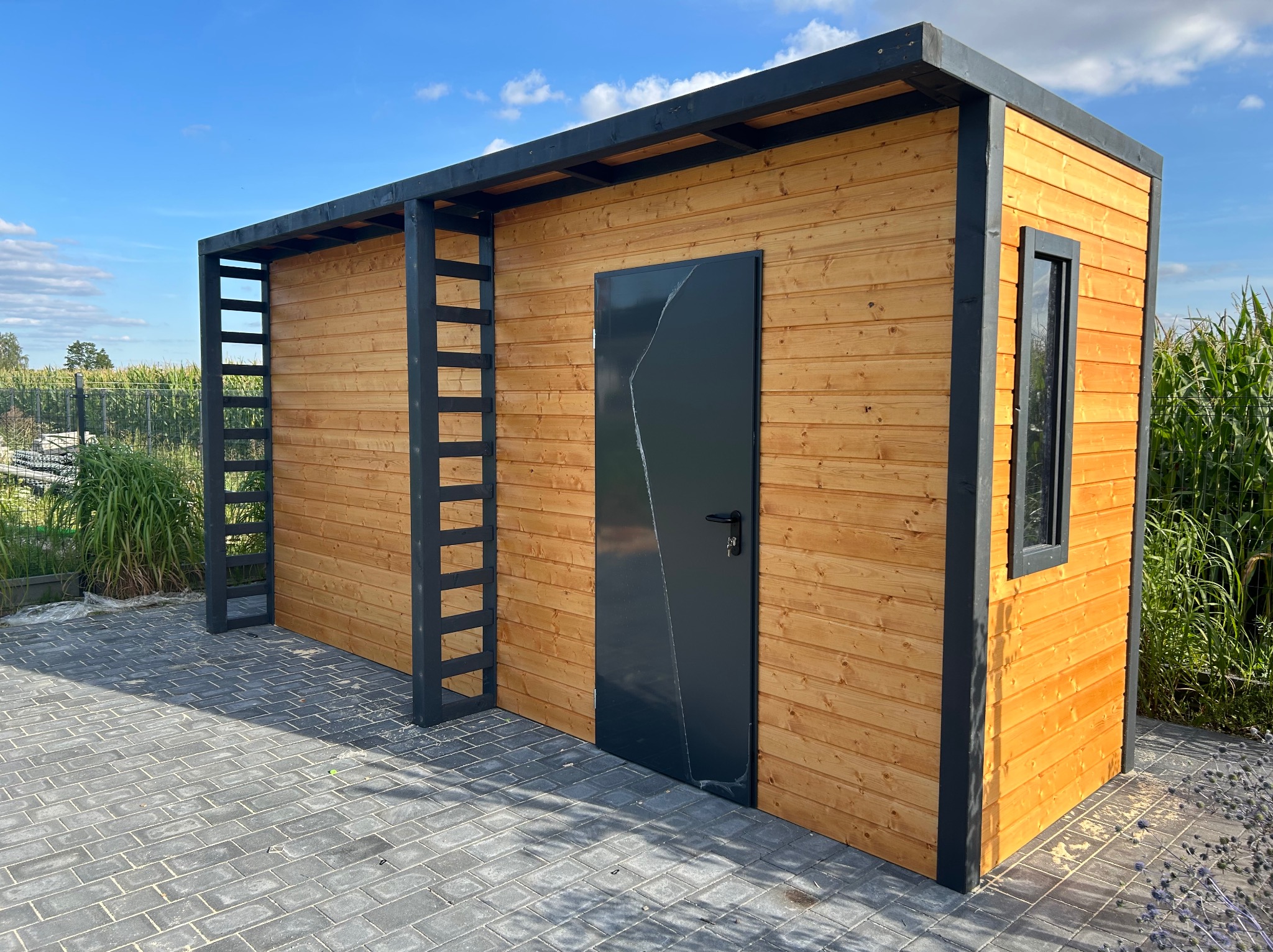 Nowoczesna, drewniana altana z czarnymi elementami konstrukcyjnymi, w tle widoczne pole kukurydzy.