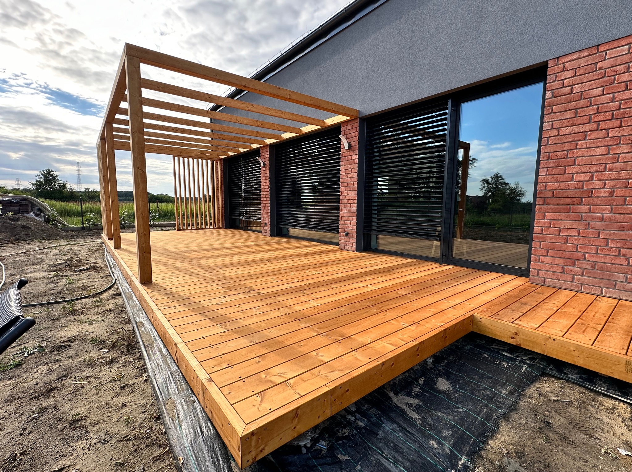 Nowoczesny taras z jasnego drewna przylegający do domu z ceglaną elewacją i szarą ścianą, z drewnianą pergolą w trakcie budowy, widoczne czarne okna z żaluzjami.