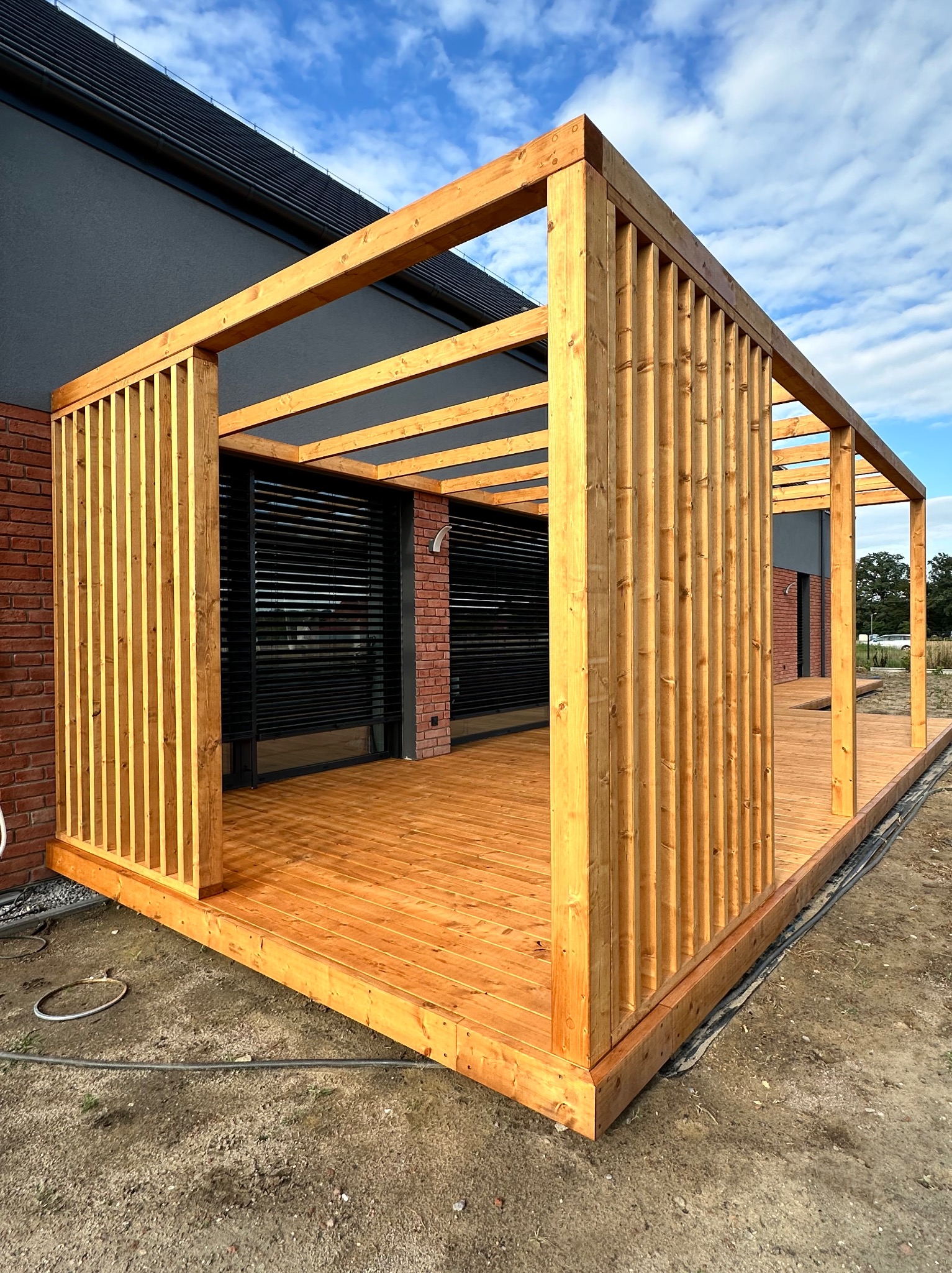 Nowoczesna, drewniana konstrukcja z ażurowymi ściankami, tworząca zadaszony taras przy budynku z ceglaną elewacją i czarnymi roletami okiennymi; widoczne elementy podłączeń elektrycznych na ziemi.