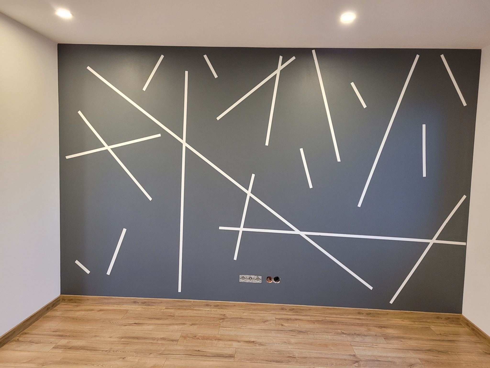 Szara ściana z abstrakcyjnym wzorem z białych linii, drewnopodobna podłoga, gniazdka elektryczne widoczne na ścianie.