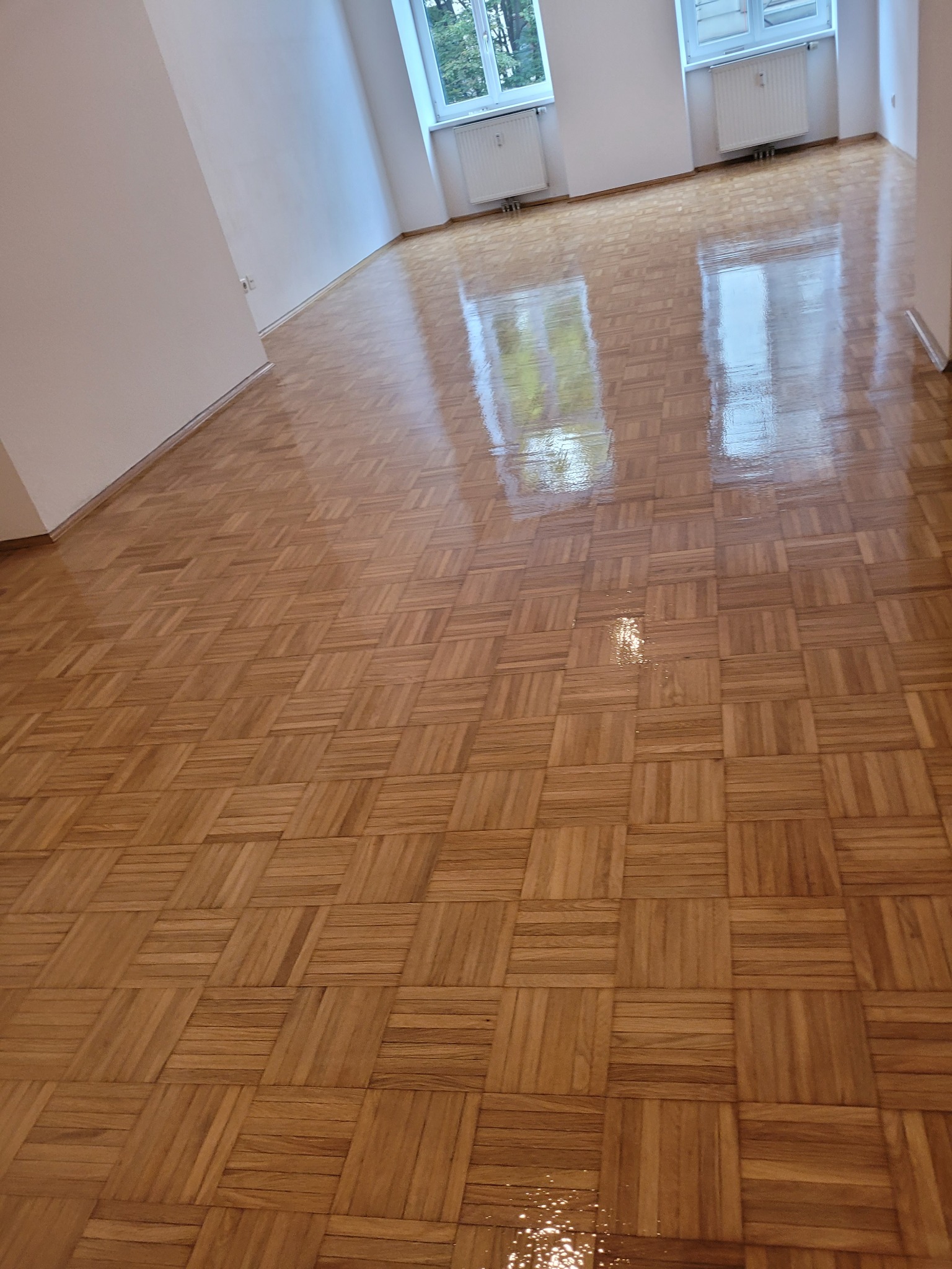 Błyszczący parkiet ułożony we wzór szachownicy, widoczne refleksy światła od okien i lamp, świeżo po nałożeniu lakieru.