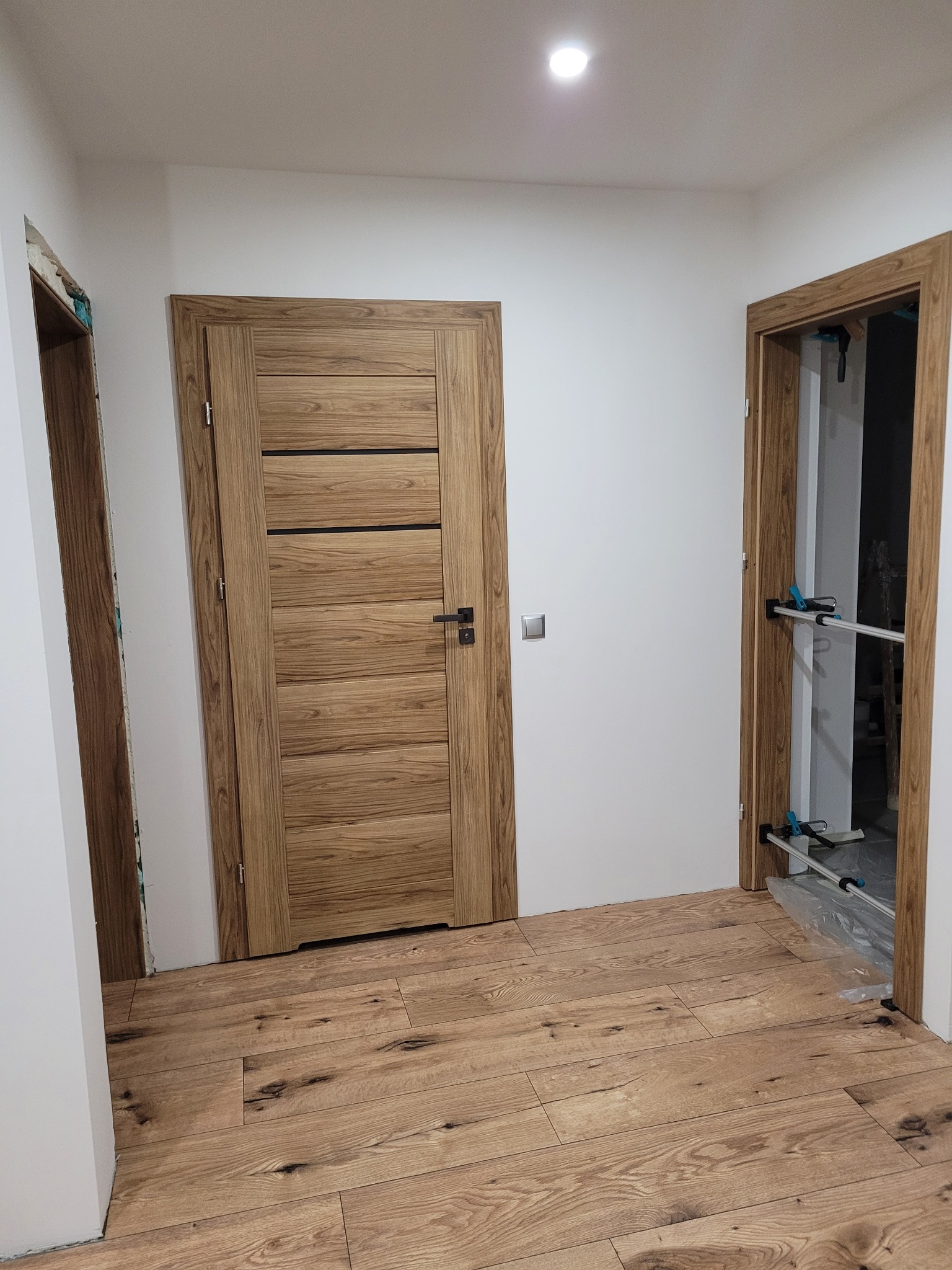 Wnętrze z drewnianą podłogą i dwiema ościeżnicami z zamontowanymi drzwiami, jedna z nich z widocznymi zaciskami stolarskimi i folią ochronną.
