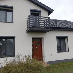 Sebo-Stal - Nowoczesny dom z balkonem z czarną, poziomą balustradą. Elewacja w jasnym odcieniu, ciemna stolarka okienna i drzwi wejściowe z dekoracyjnym przeszkleniem.