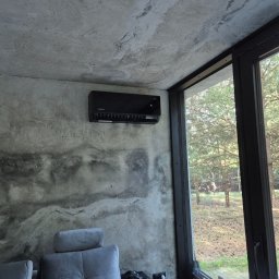 DM5 Daniel Mazuruk - Czarny klimatyzator Rotenso zamontowany na betonowej ścianie w nowoczesnym wnętrzu z widokiem na las przez duże okno.