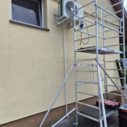 Biała klimatyzacja zamontowana na elewacji domu, widoczna z rusztowania. Przewody prowadzone wzdłuż ściany. Widoczny fragment okna i fragment dachu.