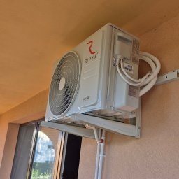 DM5 Daniel Mazuruk - Klimatyzator Rotenso Ukura Series zamontowany na balkonie, widoczne białe rurki instalacyjne i element montażowy. Beżowa ściana budynku w tle.