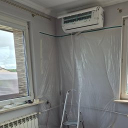 DM5 Daniel Mazuruk - Montaż klimatyzacji w pokoju zabezpieczonym folią malarską. Widoczna drabina, grzejnik i okno. Klimatyzator biały, zamontowany pod sufitem.