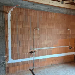 Instalacja elektryczna w budynku w trakcie budowy: przewody w ścianach z cegły ceramicznej, zabezpieczone pianką montażową, widoczne kable i puszki elektryczne.
