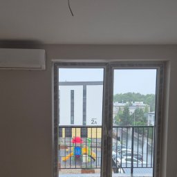 DM5 Daniel Mazuruk - Biały klimatyzator ścienny w jasnym pokoju z widokiem na plac zabaw przez okno balkonowe. Widoczne ramy okienne z logo producenta.