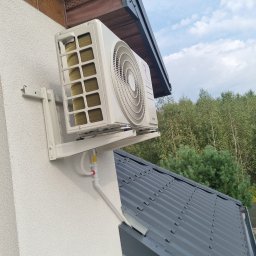 DM5 Daniel Mazuruk - Biały klimatyzator na elewacji domu, widok z dołu, z rurami odprowadzającymi skropliny. W tle dach i zieleń drzew.