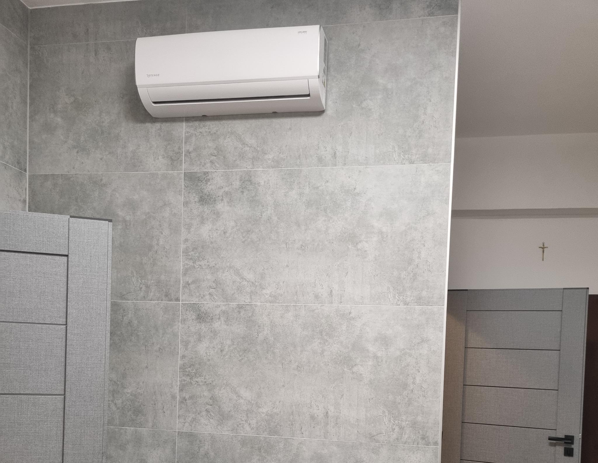 Biała klimatyzacja marki Rotenso zamontowana na szarej ścianie z płytek imitujących beton, widoczny fragment szarej zabudowy meblowej i drzwi w tle.