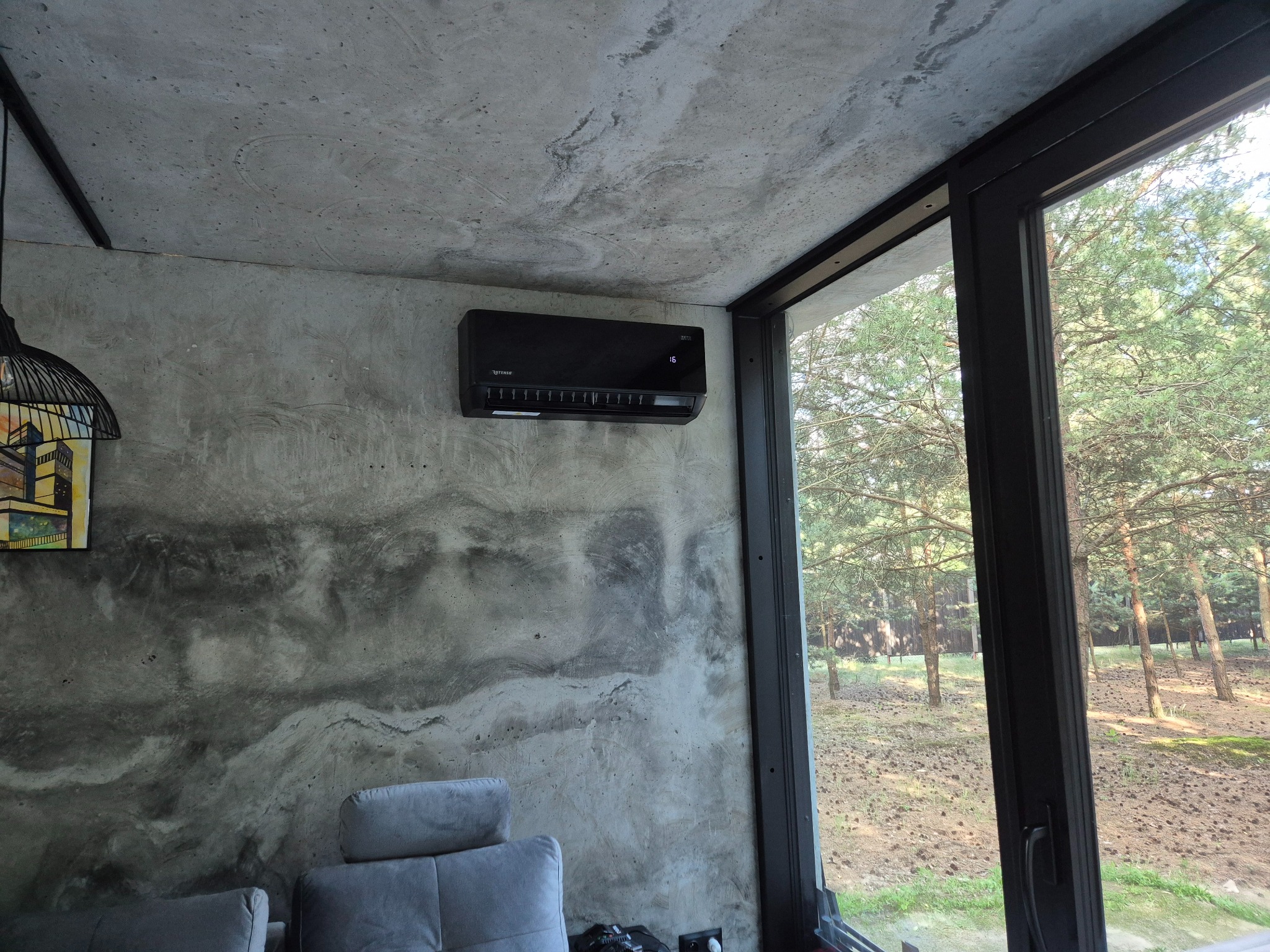 Czarny klimatyzator Rotenso zamontowany na betonowej ścianie w nowoczesnym wnętrzu z widokiem na las przez duże okno.
