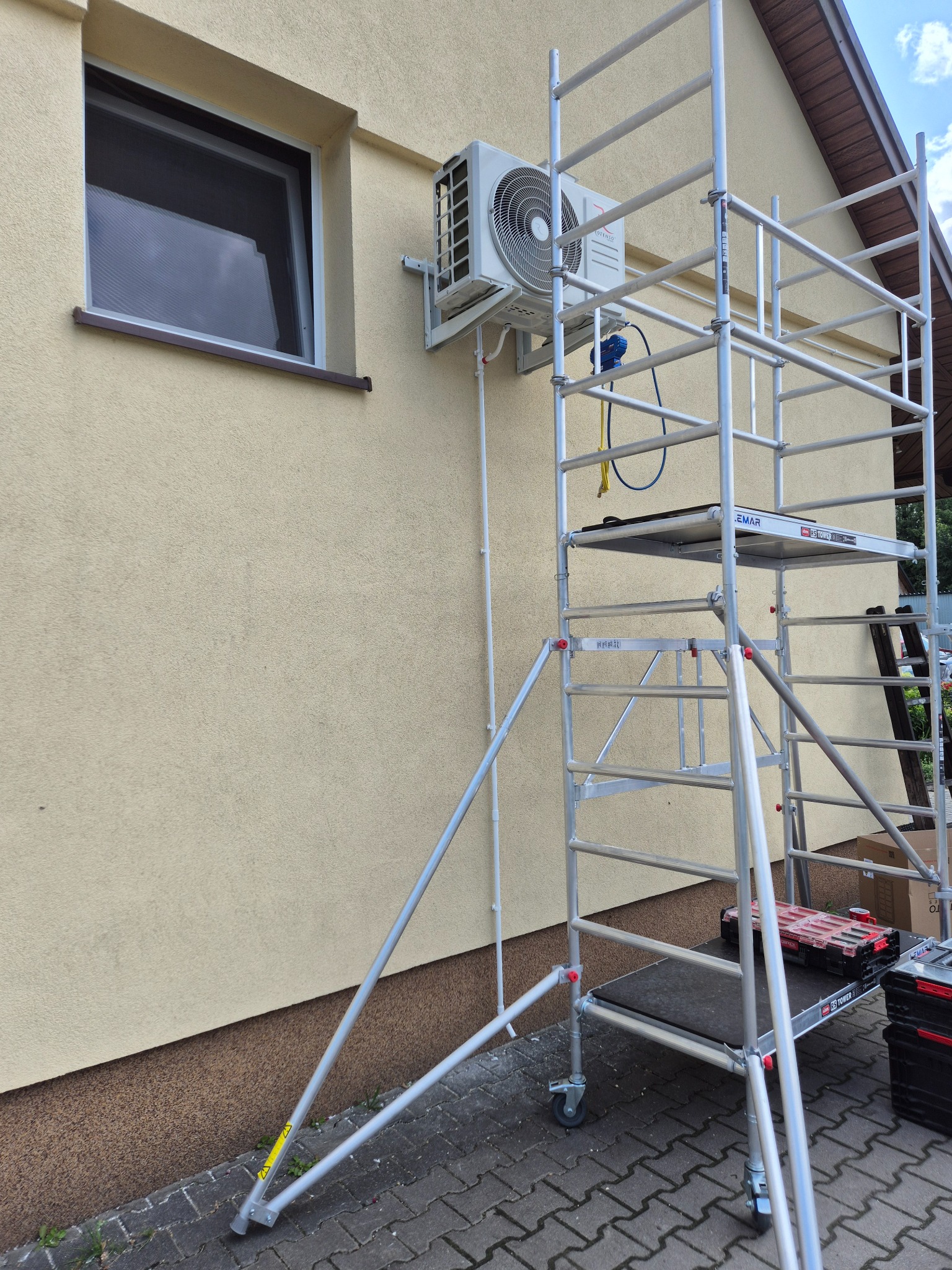 Biała klimatyzacja zamontowana na elewacji domu, widoczna z rusztowania. Przewody prowadzone wzdłuż ściany. Widoczny fragment okna i fragment dachu.