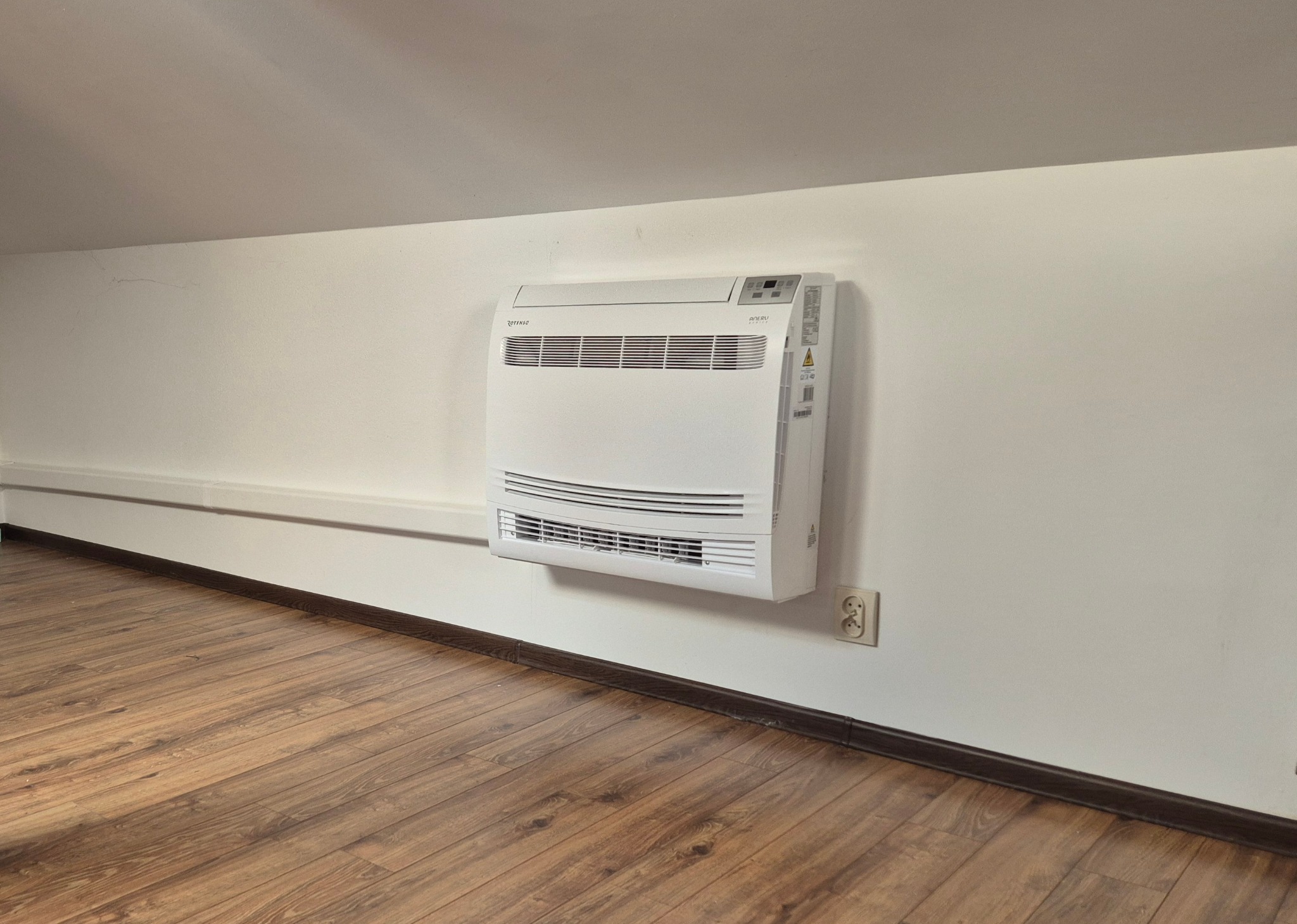 Biały, wiszący klimatyzator Rotenso AERO usytuowany na ścianie w pomieszczeniu z panelami podłogowymi. Widoczny fragment sufitu i listwa przypodłogowa.