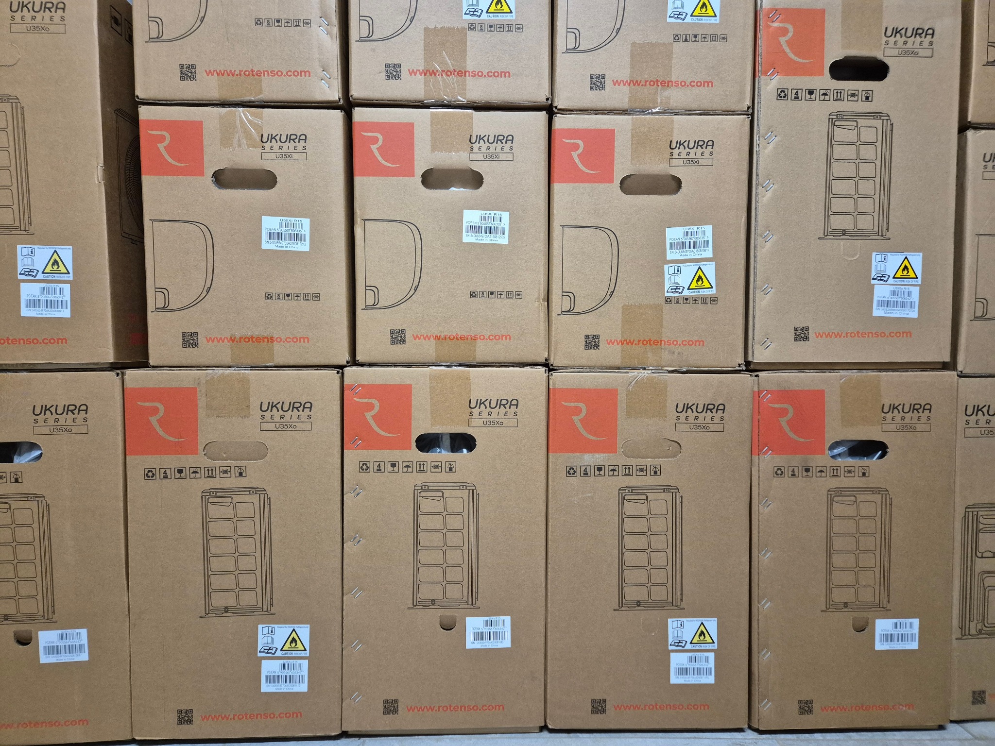 Stos kartonowych pudeł z nadrukowanymi schematami klimatyzatorów Ukura Series U35Xo i U35Xi, ułożonych jeden na drugim, z widocznymi etykietami ostrzegawczymi i logo producenta.