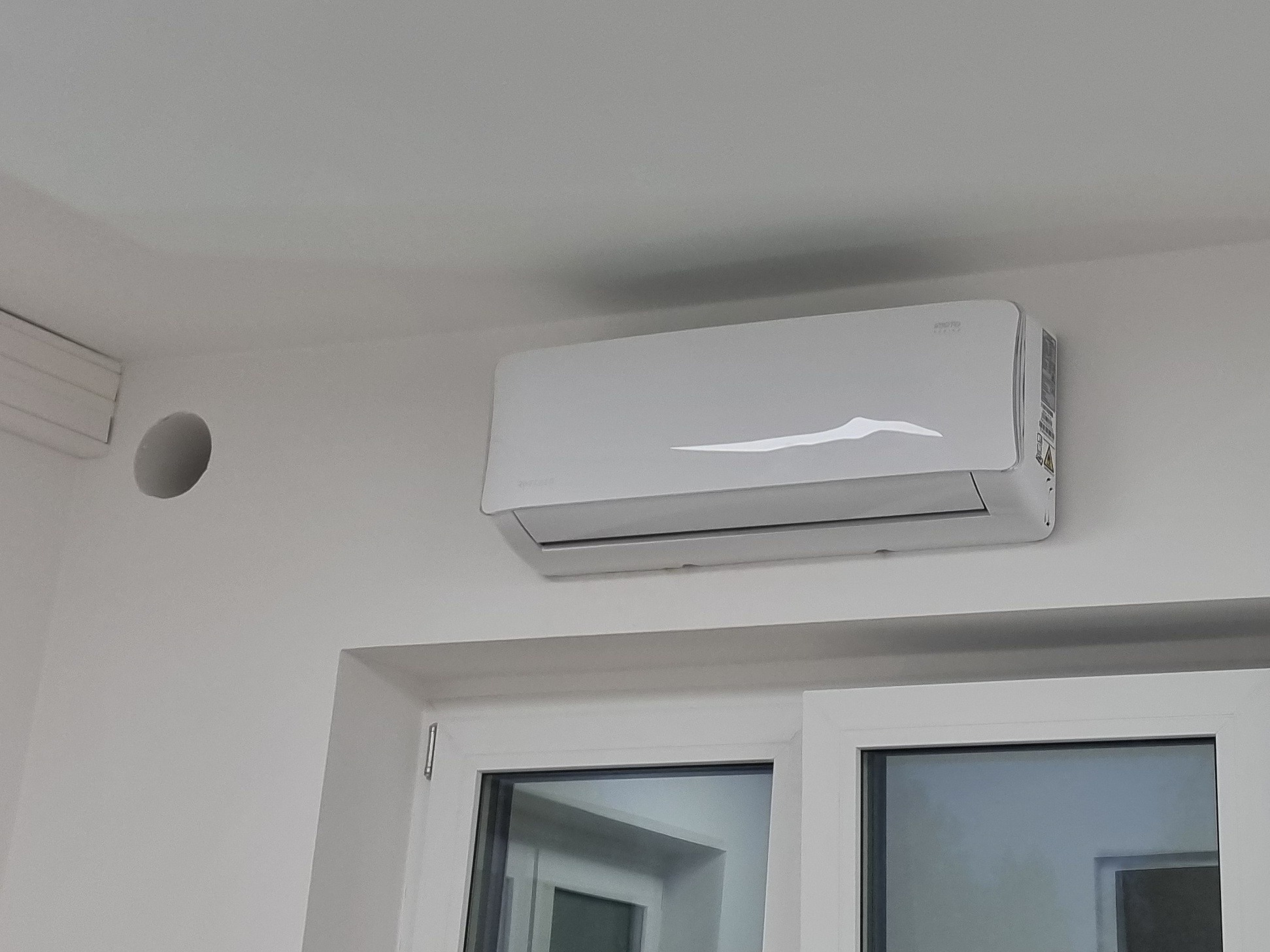 Biała klimatyzacja ścienna w jasnym wnętrzu, widoczna nad oknem. Na ścianie okrągły otwór wentylacyjny. Minimalistyczny design, świeżo zamontowane urządzenie.