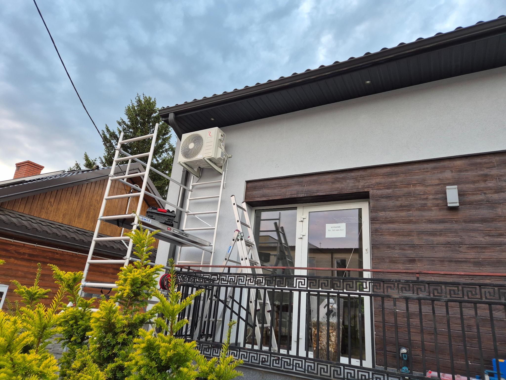 Montaż klimatyzatora na elewacji domu. Widoczne drabiny, narzędzia i jednostka zewnętrzna klimatyzacji przy oknie. Tło stanowi pochmurne niebo.