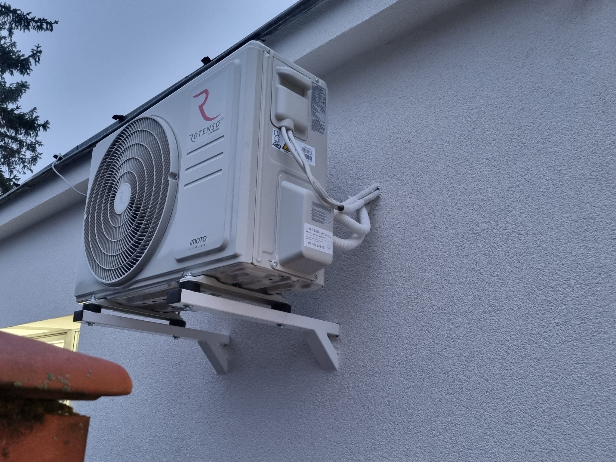 Zamontowana na zewnątrz, biała jednostka klimatyzacji Rotenso Imoto Series na wspornikach. Widoczne przewody i fragment dachu z dachówką. Teksturowana ściana budynku.