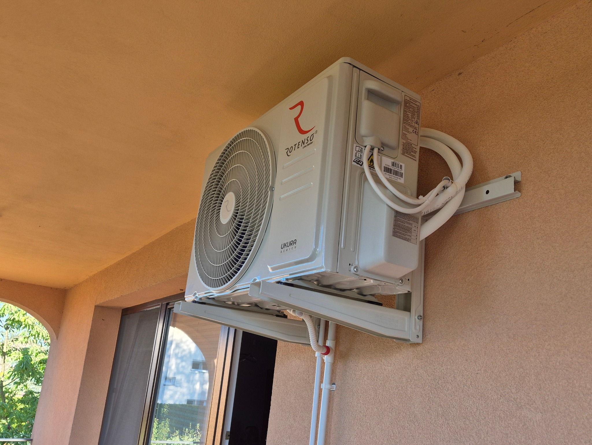 Klimatyzator Rotenso Ukura Series zamontowany na balkonie, widoczne białe rurki instalacyjne i element montażowy. Beżowa ściana budynku w tle.
