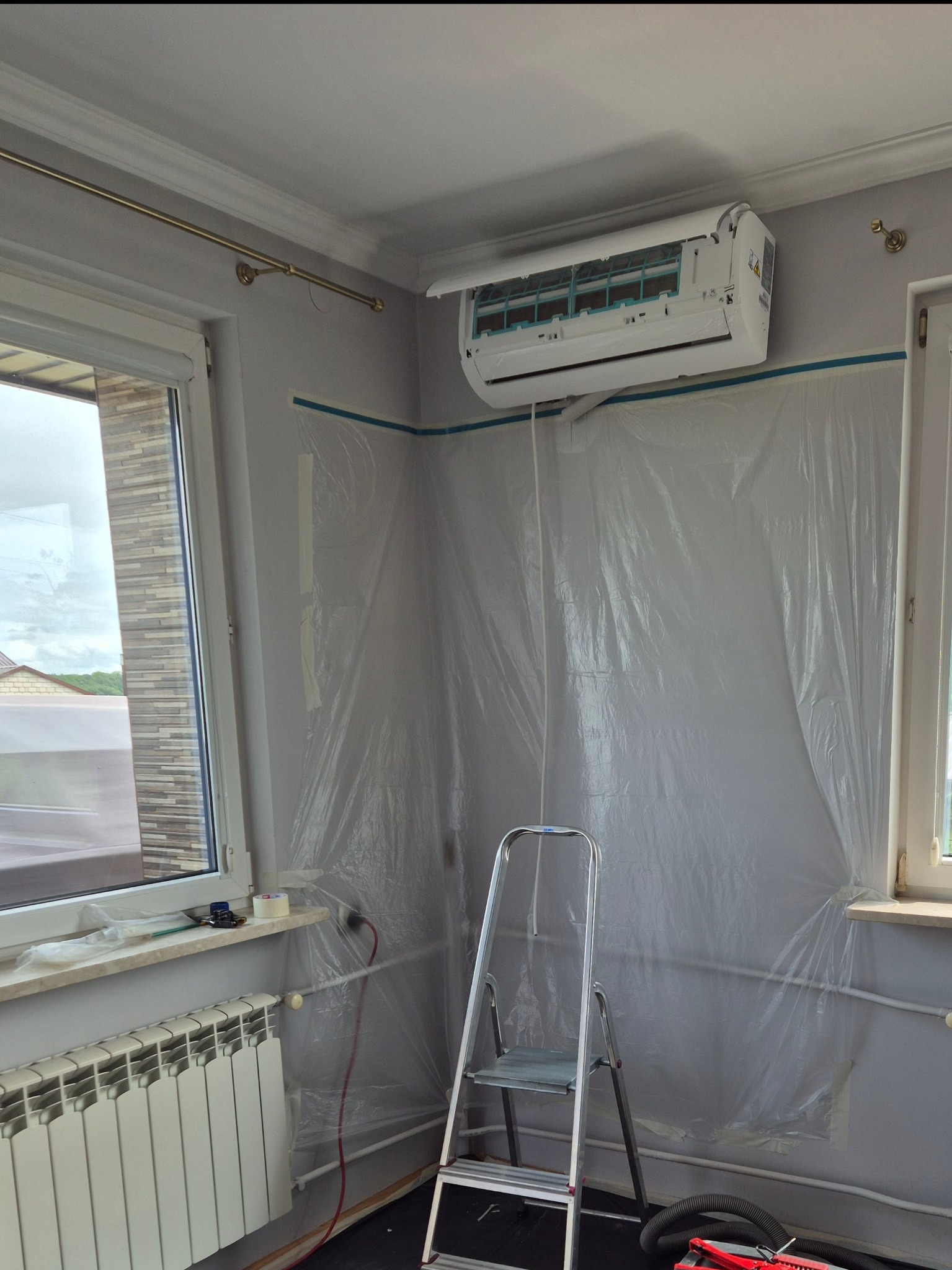 Montaż klimatyzacji w pokoju zabezpieczonym folią malarską. Widoczna drabina, grzejnik i okno. Klimatyzator biały, zamontowany pod sufitem.