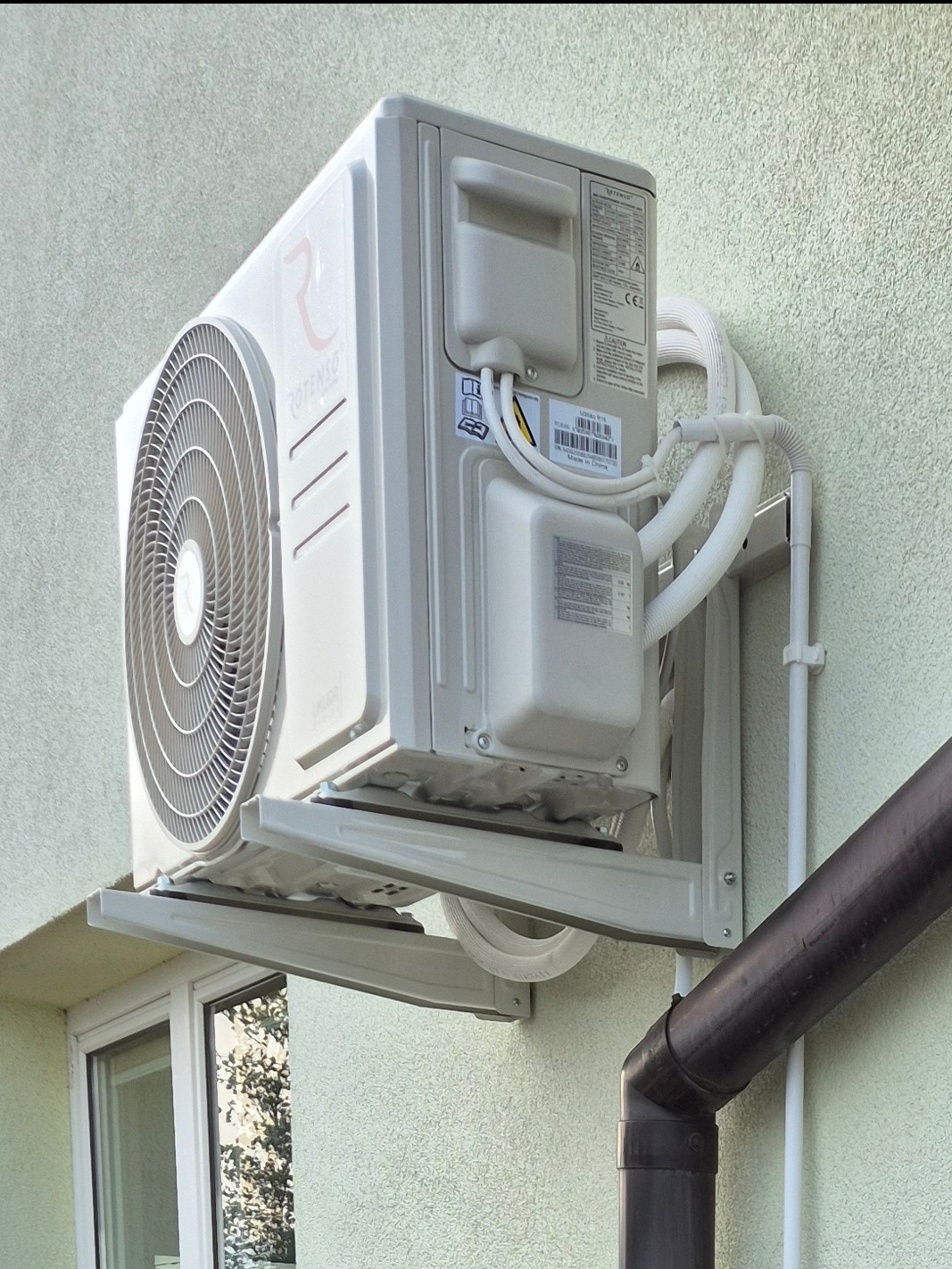 Biały klimatyzator Rotenso zamontowany na elewacji budynku. Widoczne rury odprowadzające ciepło i okno w tle. Ujęcie z dołu.