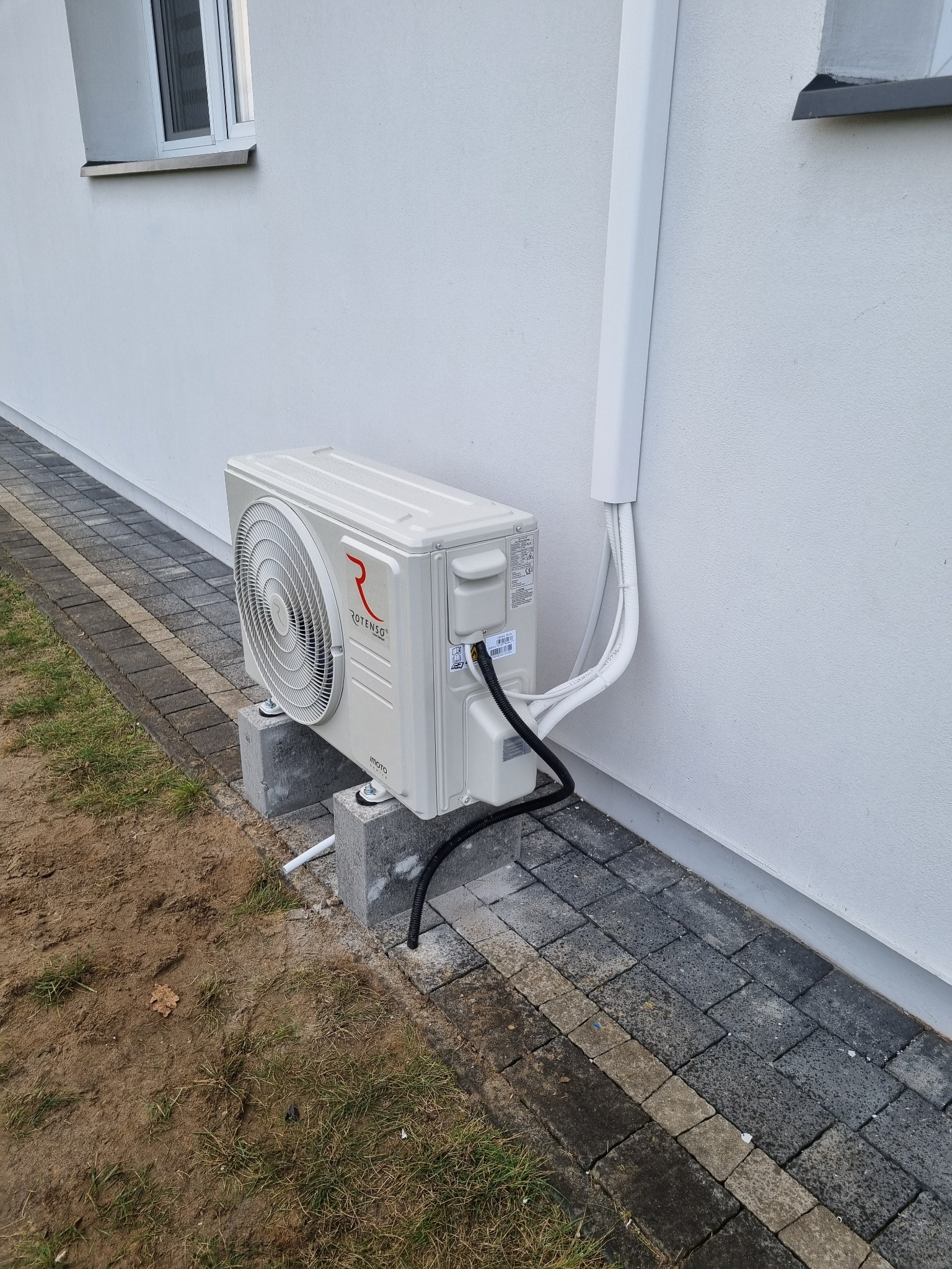 Zewnętrzna jednostka klimatyzacji Rotenso zamontowana na betonowych bloczkach przy elewacji budynku. Widoczne przewody w białej osłonie.