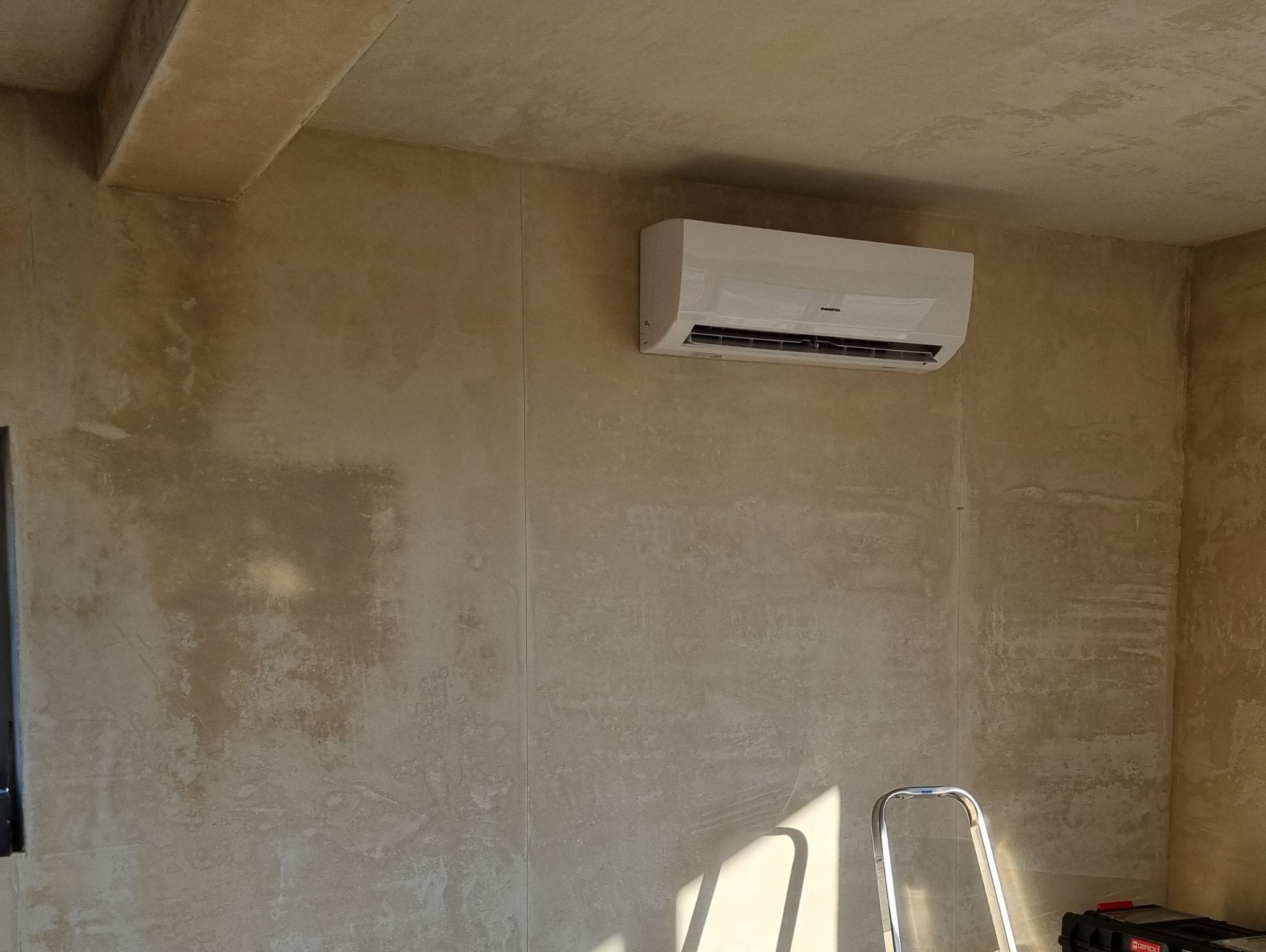 Zamontowany klimatyzator INNOVA na ścianie o fakturze betonu. Obok drabina i skrzynka narzędziowa. Widoczne światło słoneczne.