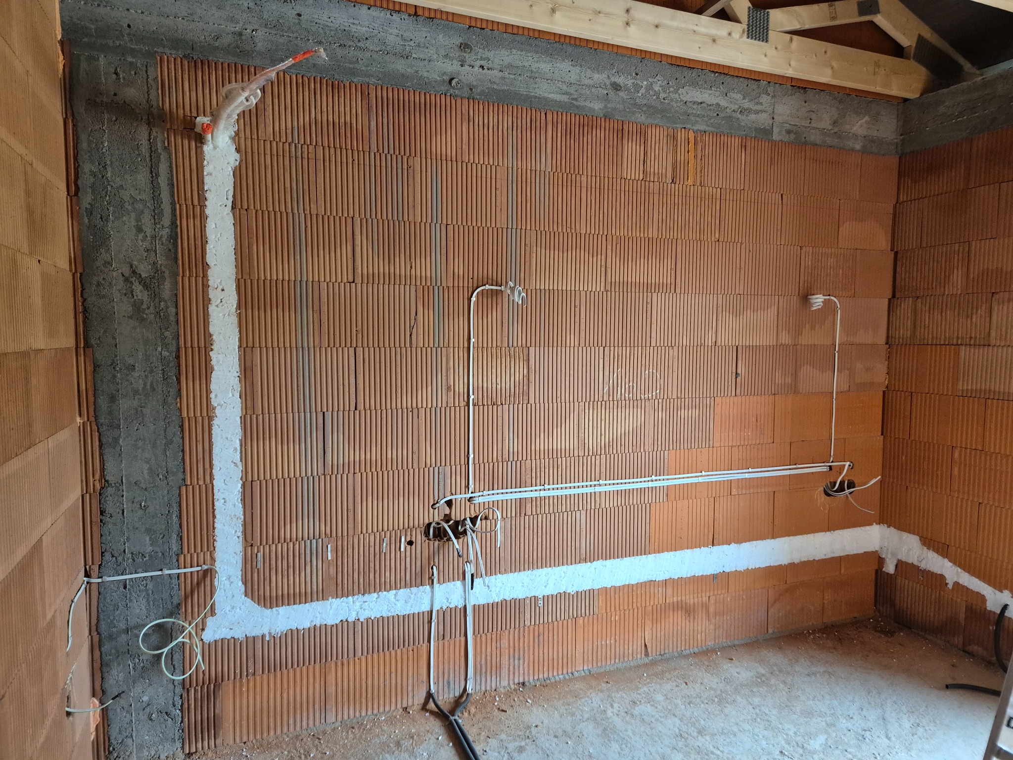 Instalacja elektryczna w budynku w trakcie budowy: przewody w ścianach z cegły ceramicznej, zabezpieczone pianką montażową, widoczne kable i puszki elektryczne.