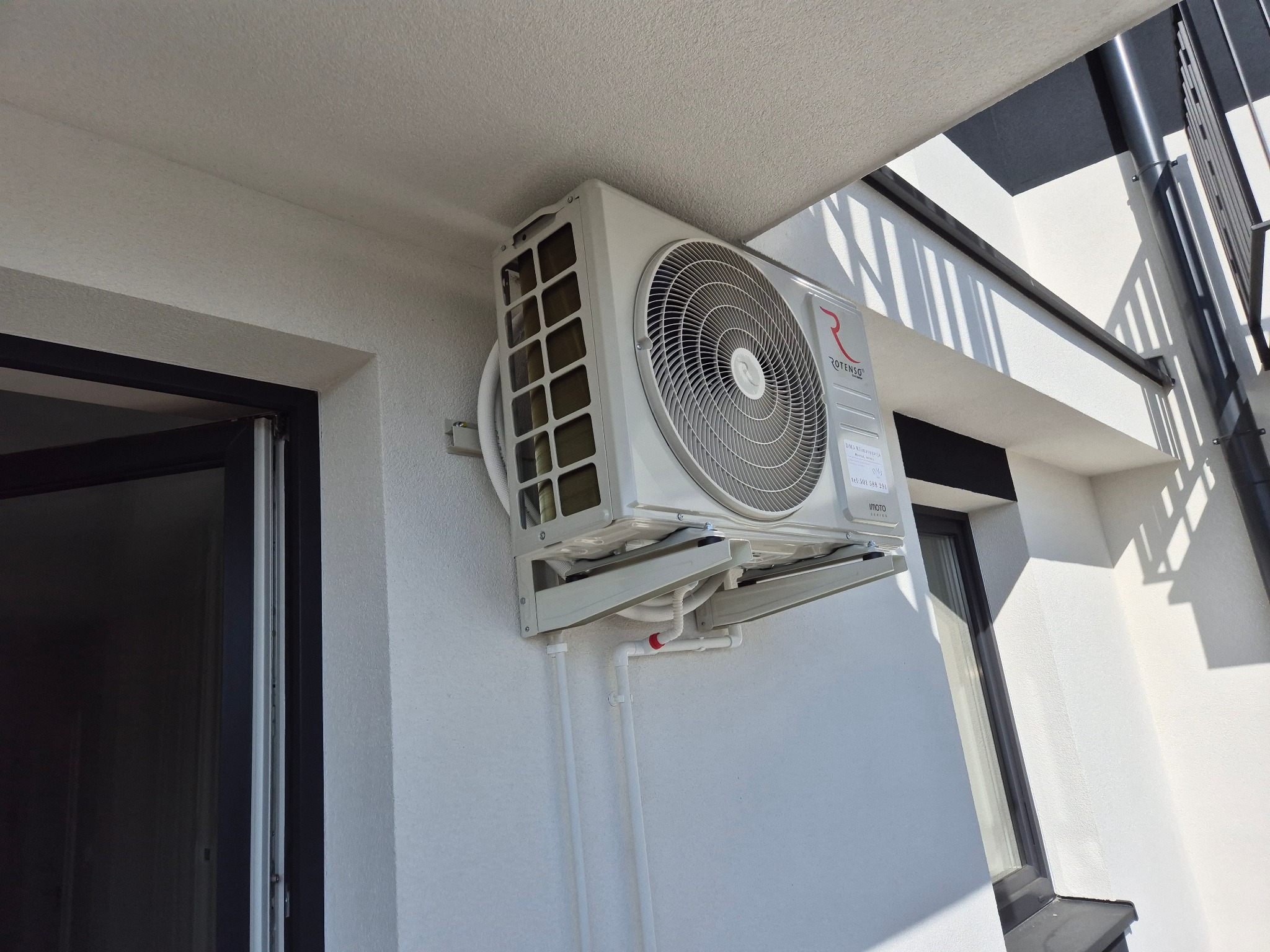 Zamontowana jednostka zewnętrzna klimatyzacji Rotenso na białej ścianie budynku. Widoczne białe rury odprowadzające skropliny. Część okna i balustrady w tle.