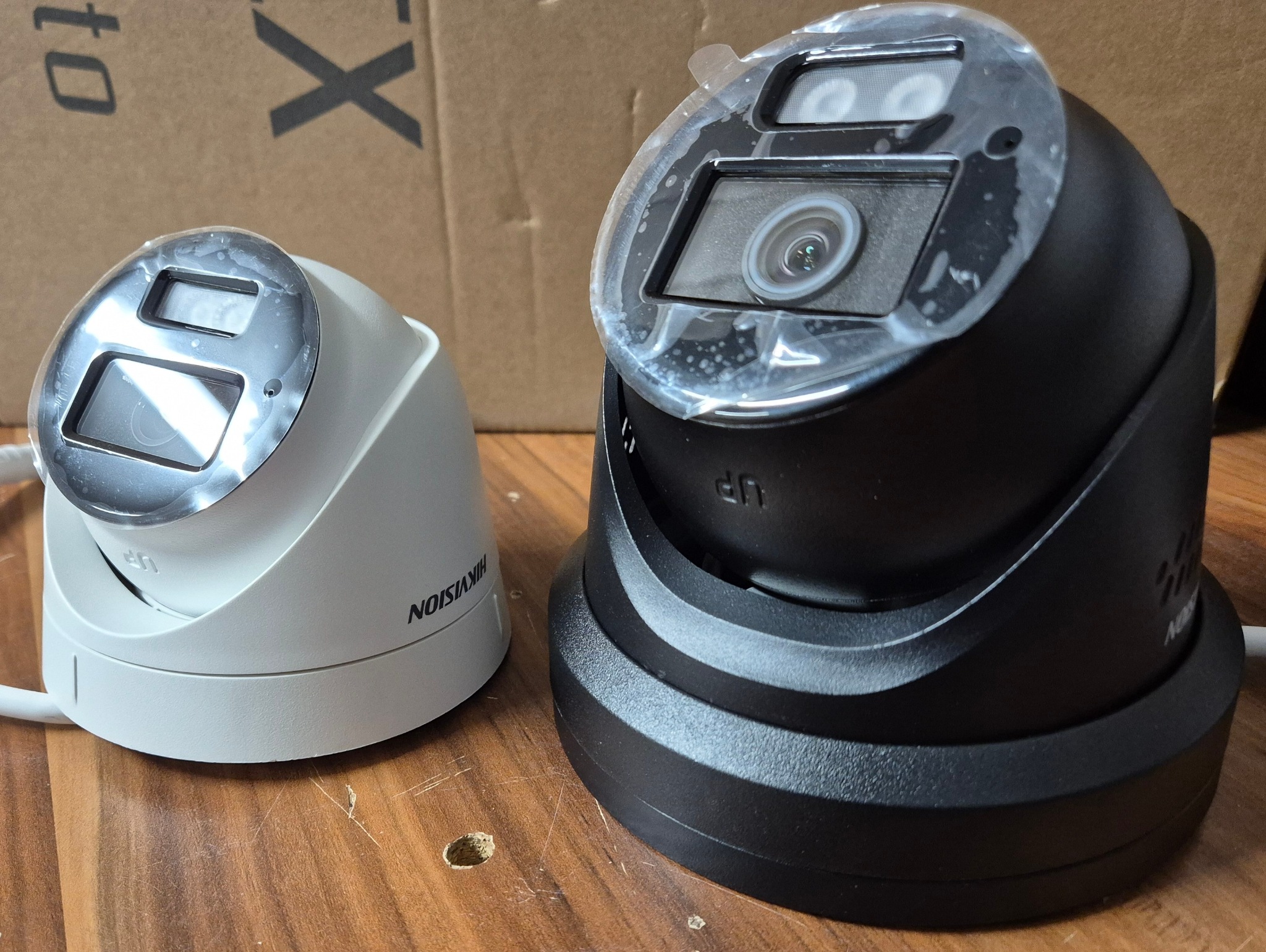 Dwie kamery kopułkowe Hikvision: biała i czarna, z folią ochronną na obiektywach, leżą na drewnianym blacie. Widoczne logo producenta i oznaczenie 'UP'.
