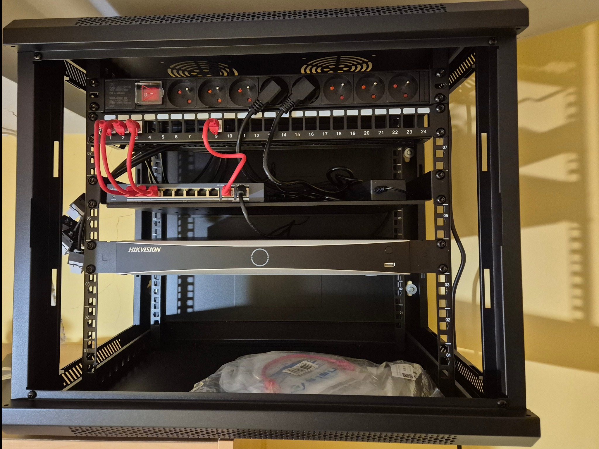 Czarna szafa rack z zamontowanym sprzętem sieciowym: patch panel, switch, rejestrator Hikvision. Widoczne kable Ethernet i zasilające. Gniazda zasilające z wyłącznikiem.