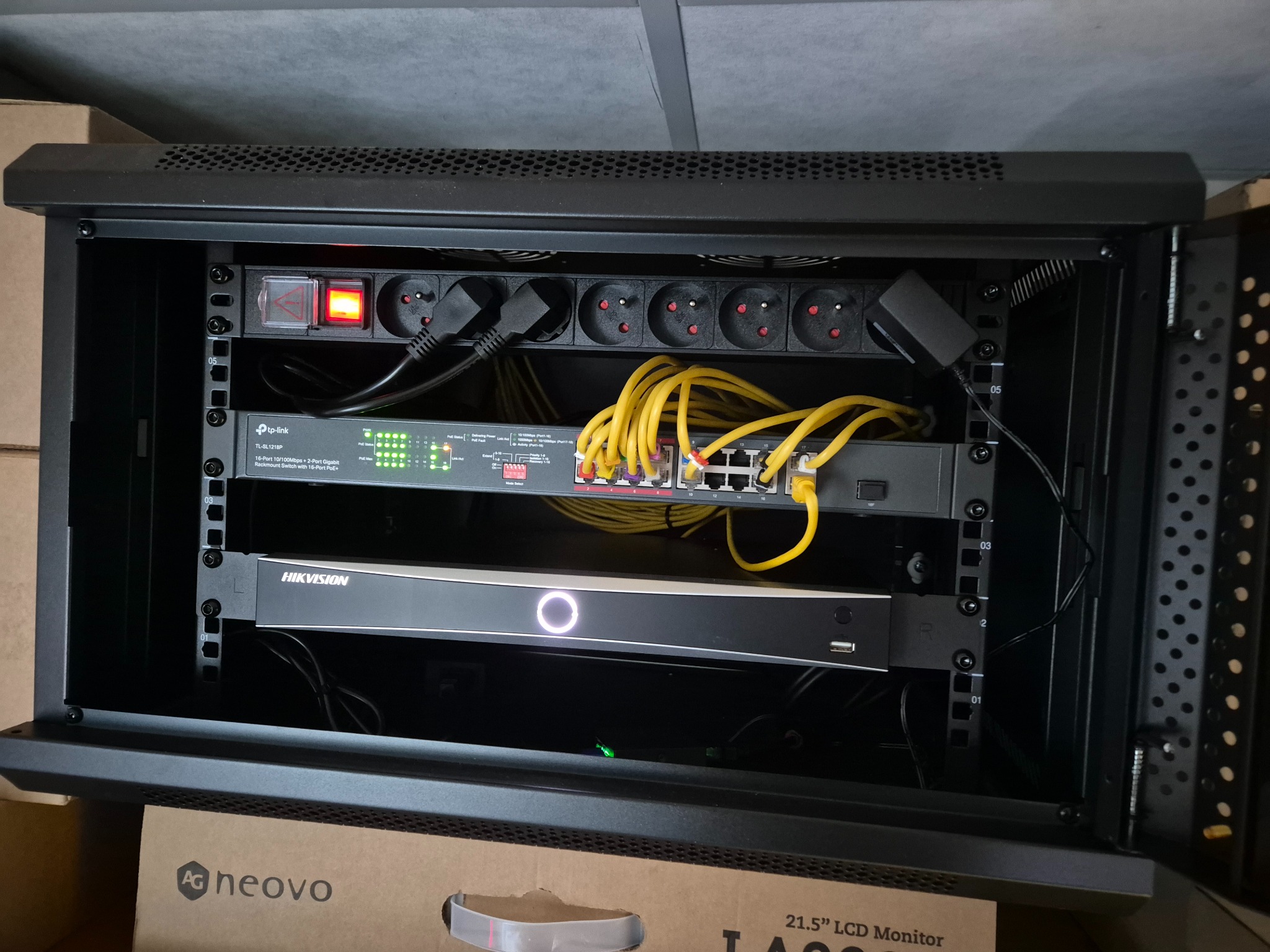 Szafa rack z zamontowanym switchem TP-Link, rejestratorem Hikvision i listwą zasilającą. Żółte kable sieciowe podłączone do switcha. Widoczne oznaczenia wysokości w szafie.