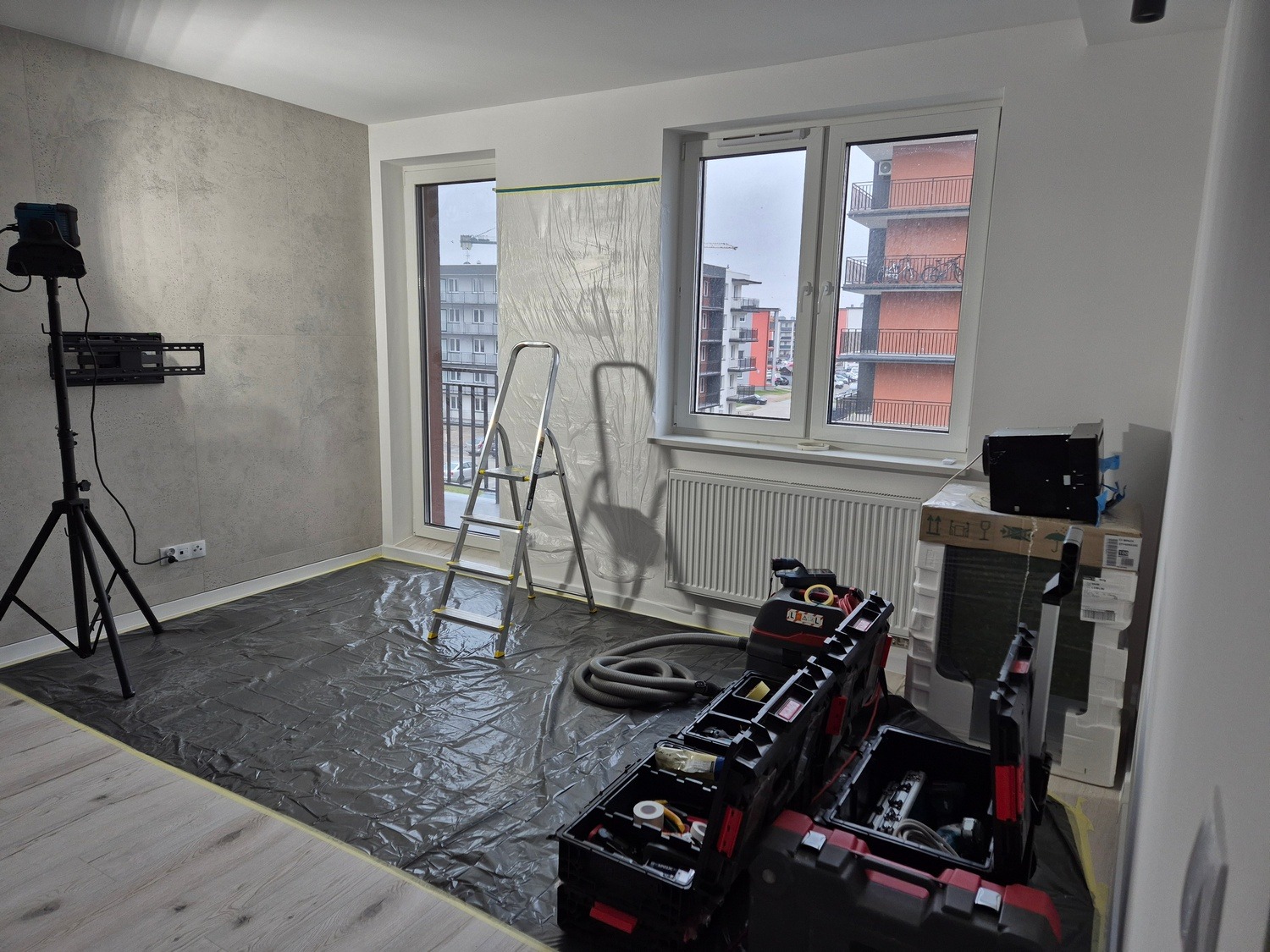 Przygotowanie do montażu klimatyzacji w mieszkaniu: zabezpieczona podłoga folią, drabina, narzędzia i agregat klimatyzacyjny przy oknie. Widok na osiedle z okna.