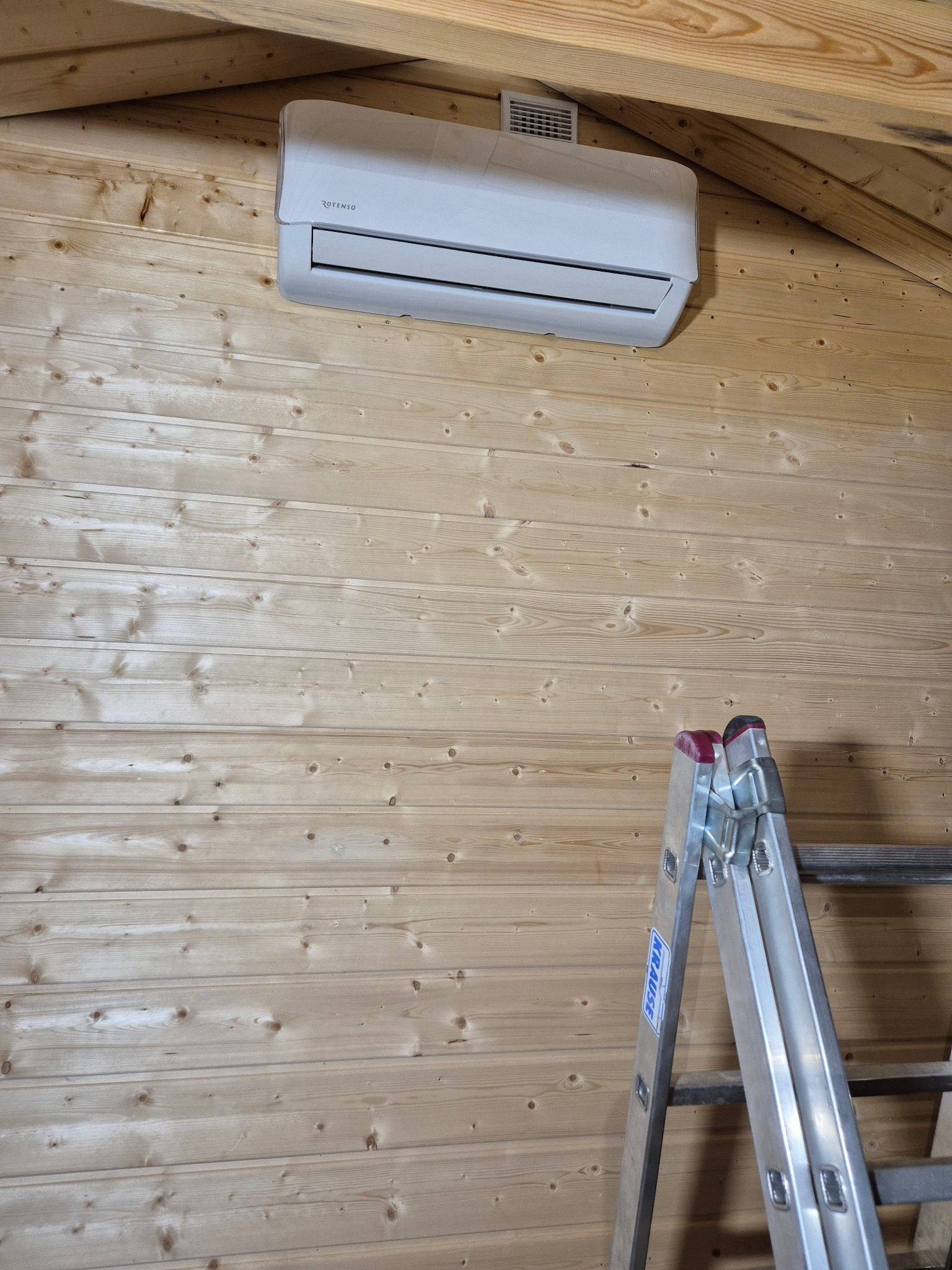 Biała klimatyzacja Rotenso zamontowana na drewnianej ścianie poddasza, widoczna drabina Krause. Ujęcie z dołu, podkreślające instalację w trudnych warunkach.