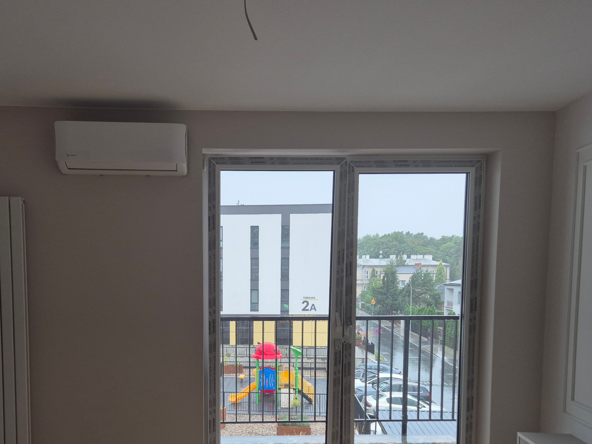 Biały klimatyzator ścienny w jasnym pokoju z widokiem na plac zabaw przez okno balkonowe. Widoczne ramy okienne z logo producenta.