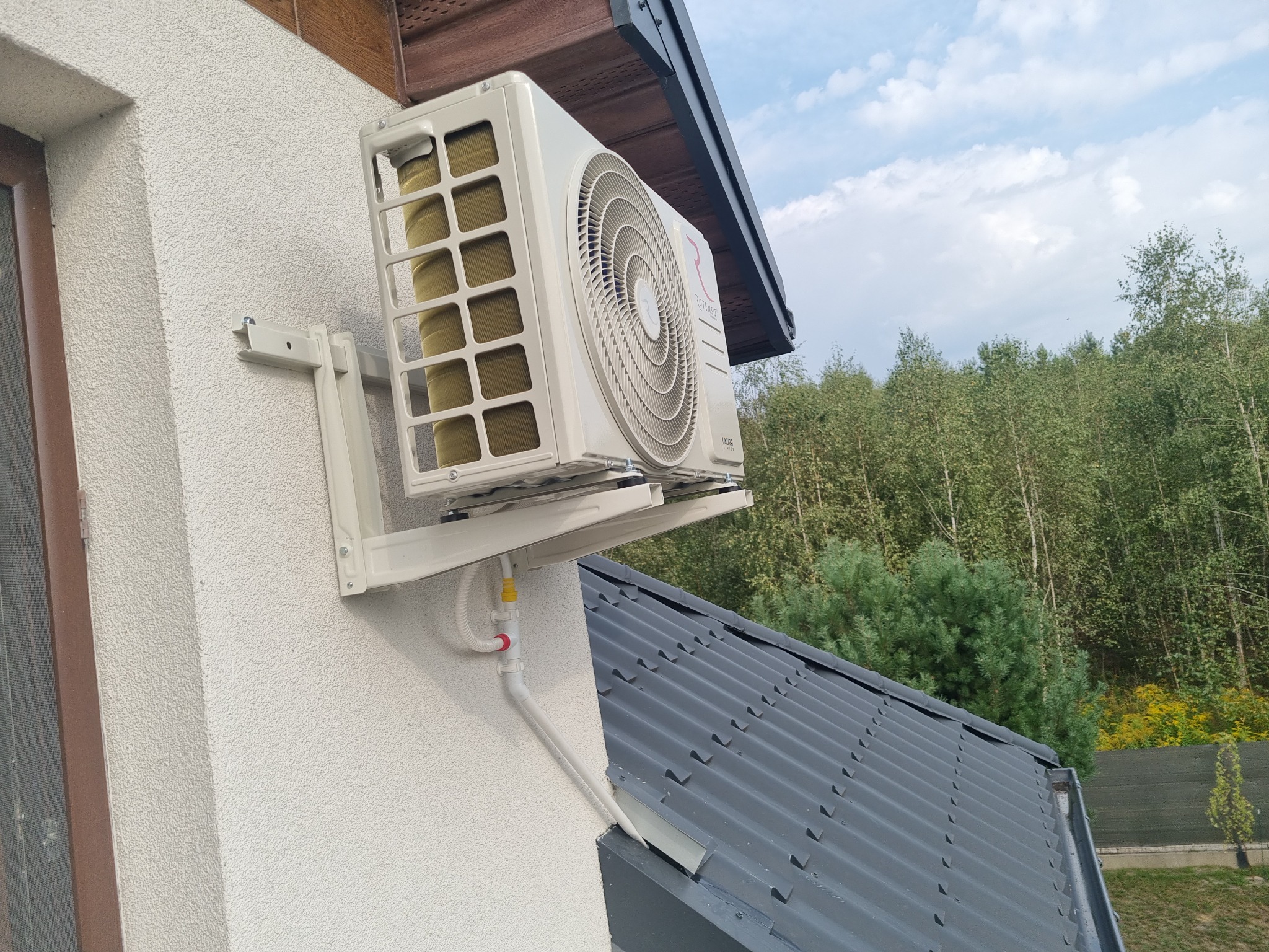 Biały klimatyzator na elewacji domu, widok z dołu, z rurami odprowadzającymi skropliny. W tle dach i zieleń drzew.