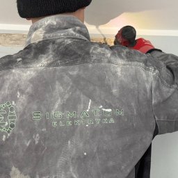 SIGMATOM SPÓŁKA Z OGRANICZONĄ ODPOWIEDZIALNOŚCIĄ - Elektryk w roboczej, poplamionej kurtce z logo firmy 'SIGMATOM Elektryka' wierci otwór w suficie za pomocą wiertarki akumulatorowej, widoczne przewody w ścianie.