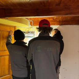 SIGMATOM SPÓŁKA Z OGRANICZONĄ ODPOWIEDZIALNOŚCIĄ - Trzech elektryków w kurtkach firmowych 'SIGMATOM Elektryka' sprawdza skrzynkę elektryczną w budynku z drewnianym sufitem i żółtą ścianą.