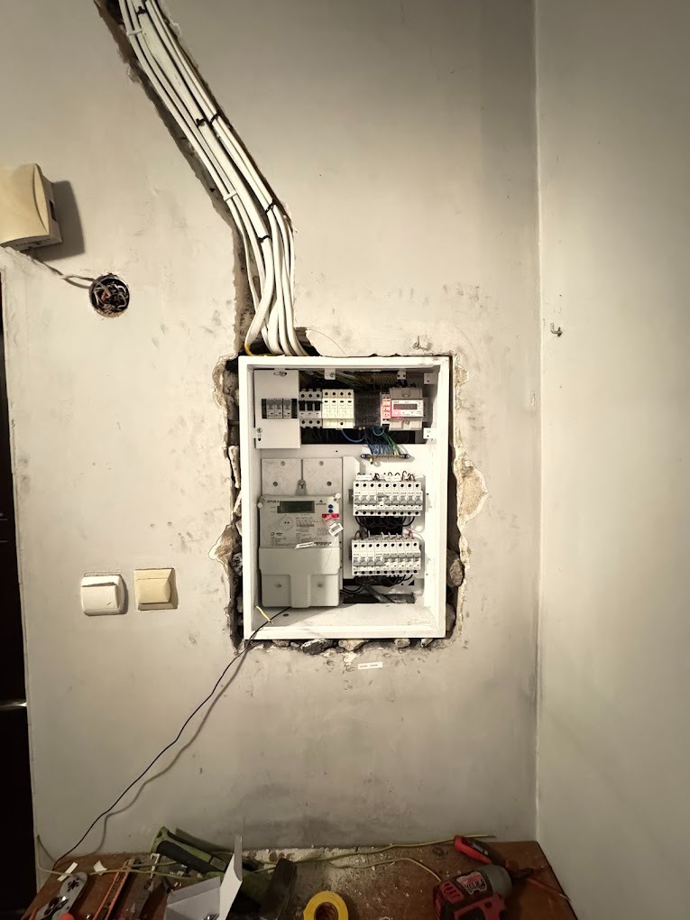 Otwarta skrzynka elektryczna z licznikiem i bezpiecznikami w surowej ścianie, widoczne przewody i narzędzia na podłodze.
