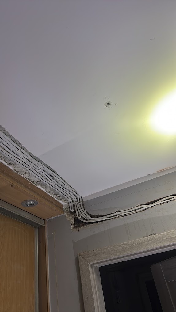 Wiązka kabli elektrycznych ułożona w bruździe pod sufitem podczas remontu, widoczna lampa halogenowa i fragment futryny drzwi.